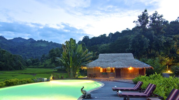 Zona de piscina con sillones reclinables, una estructura de techo de paja y vista a un paisaje verde exuberante.