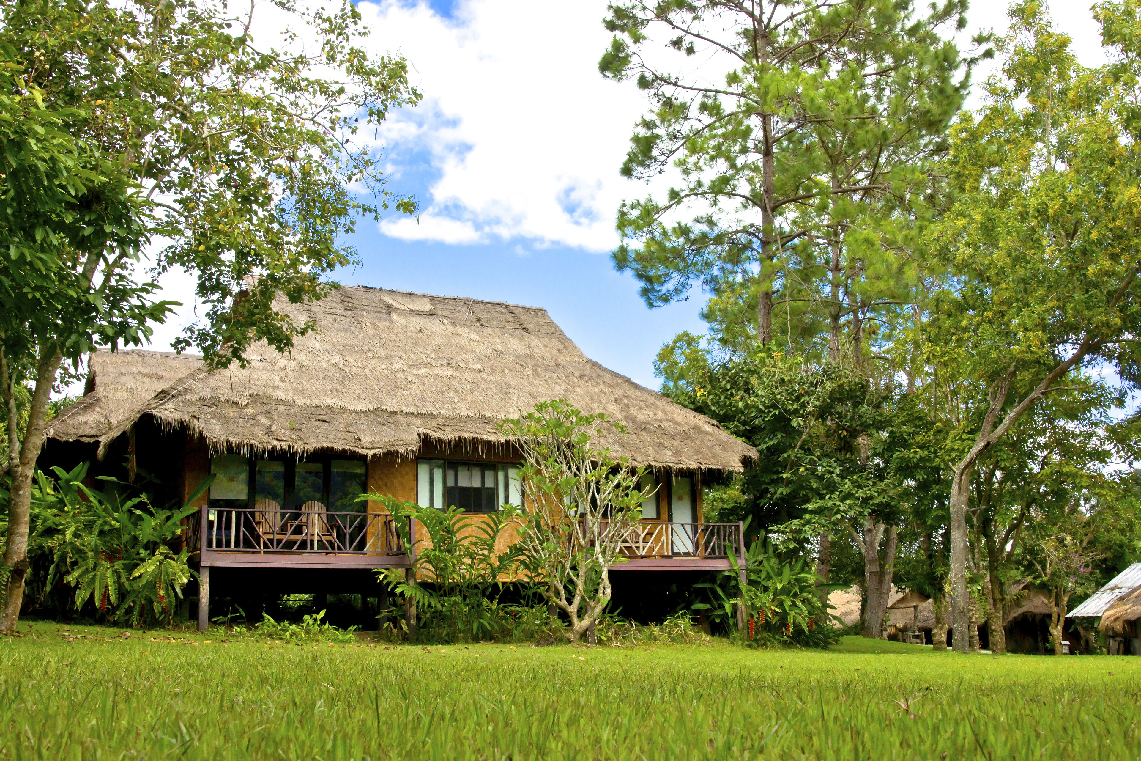 A thatched-roof bungalow with a balcony, surrounded by lush greenery.