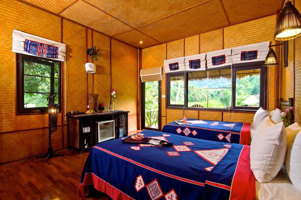 A room with two beds, wooden walls, large windows, a desk, and a fan.