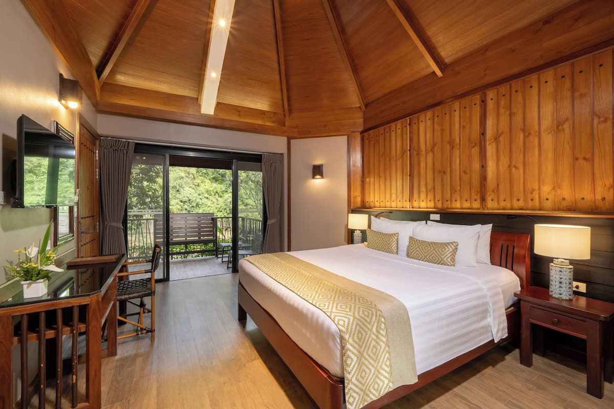 A spacious bedroom with a large bed, wooden ceiling, a desk with a chair, and a balcony with a view of greenery.