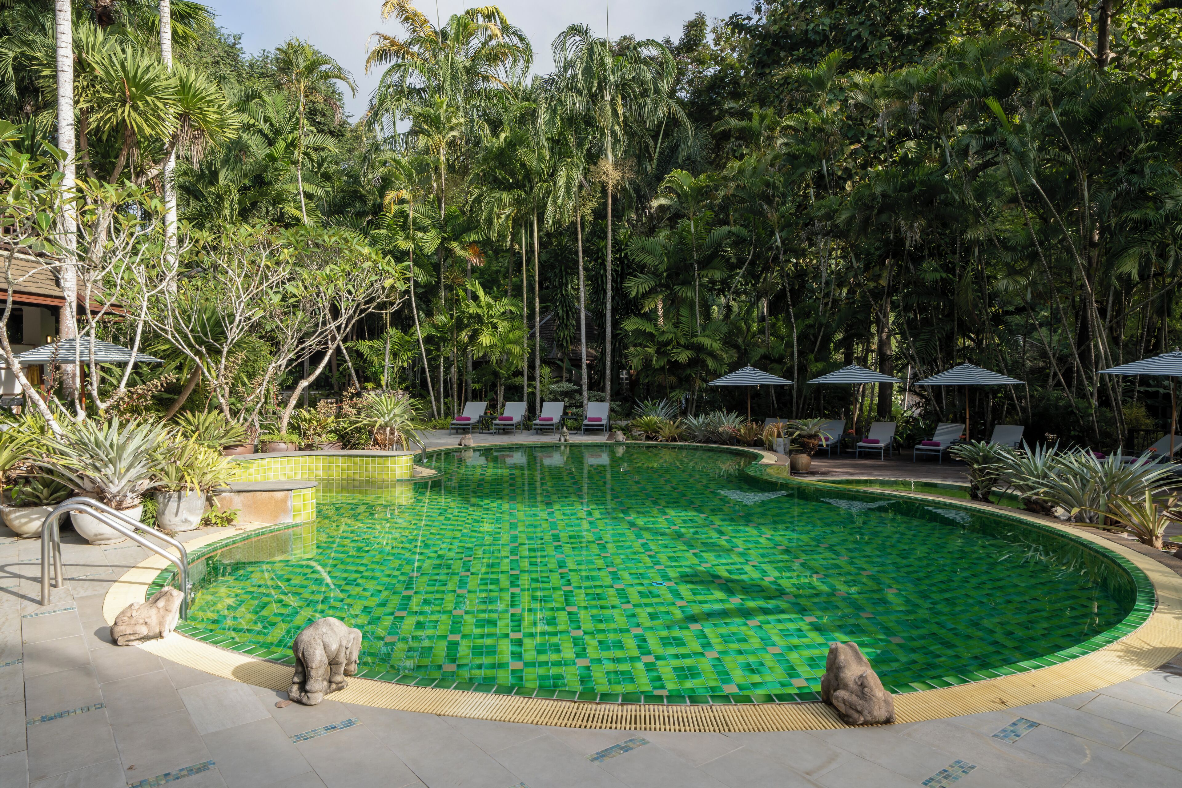 Ein Pool mit grünem Mosaikmuster, umgeben von Liegestühlen und Sonnenschirmen, mit Palmen und anderer Vegetation im Hintergrund.