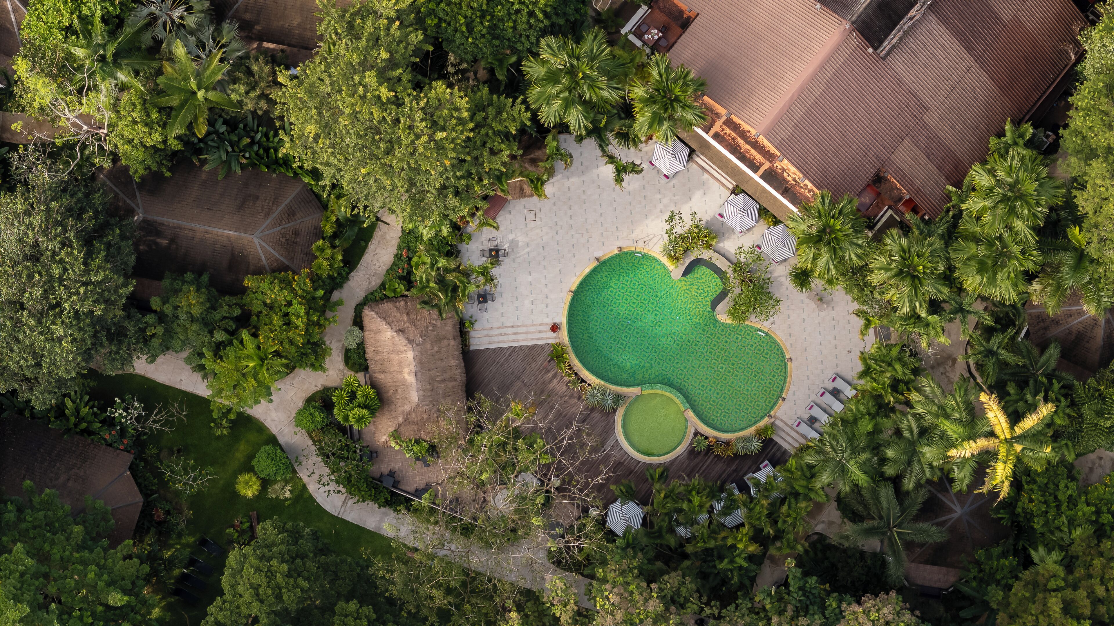 An aerial view of a resort with a central swimming pool, surrounded by lush greenery and palm trees.
