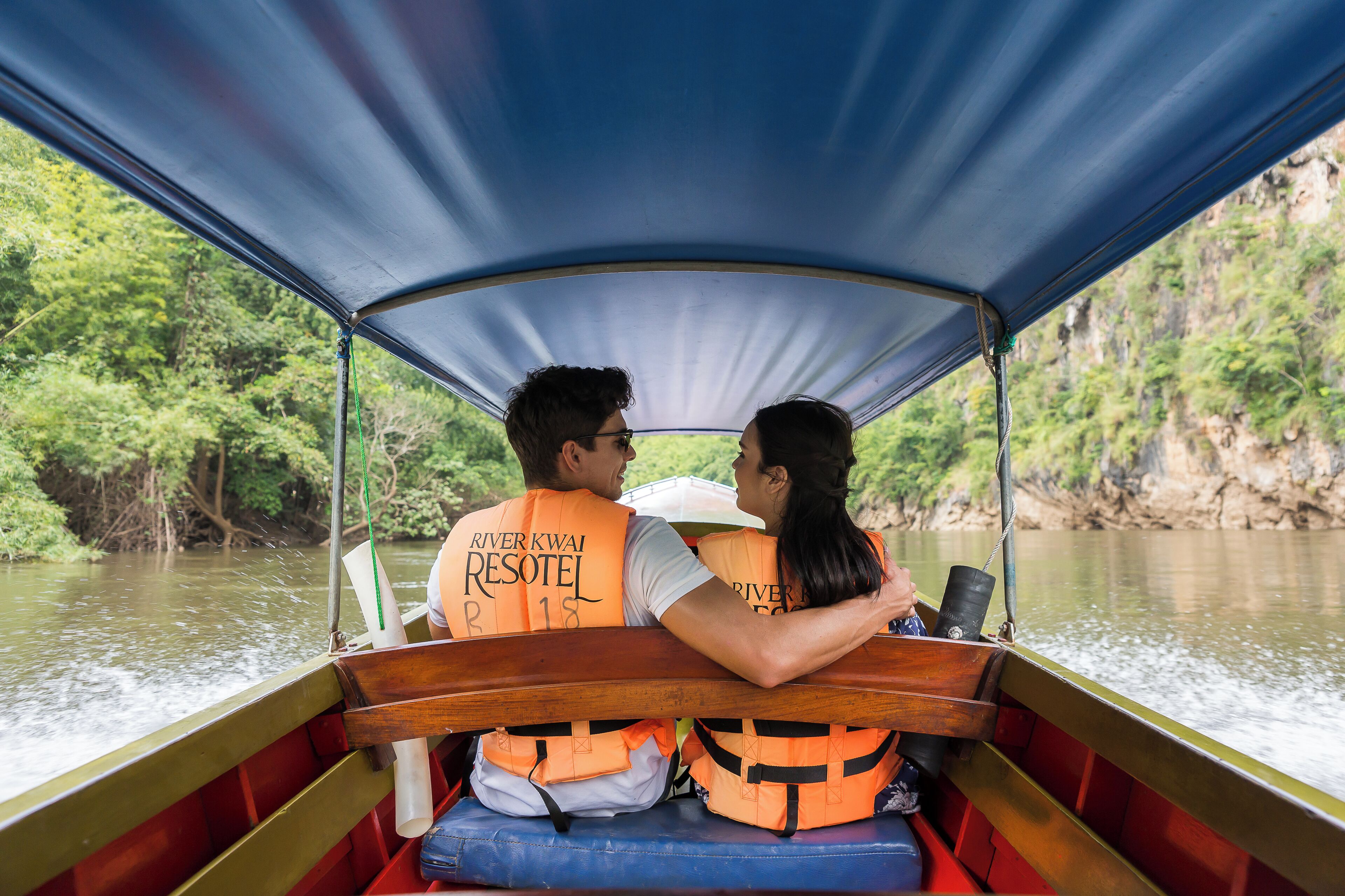A couple on a boat with a canopy, wearing life jackets, and a river view.