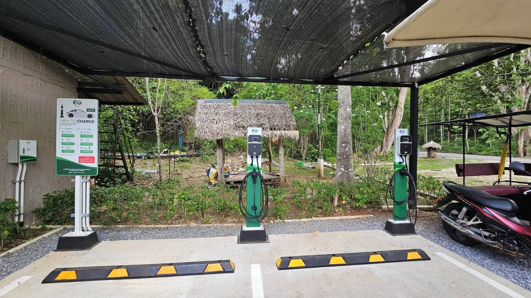 A covered parking area with electric vehicle charging stations, a thatched-roof structure, and a motorcycle parked to the side.