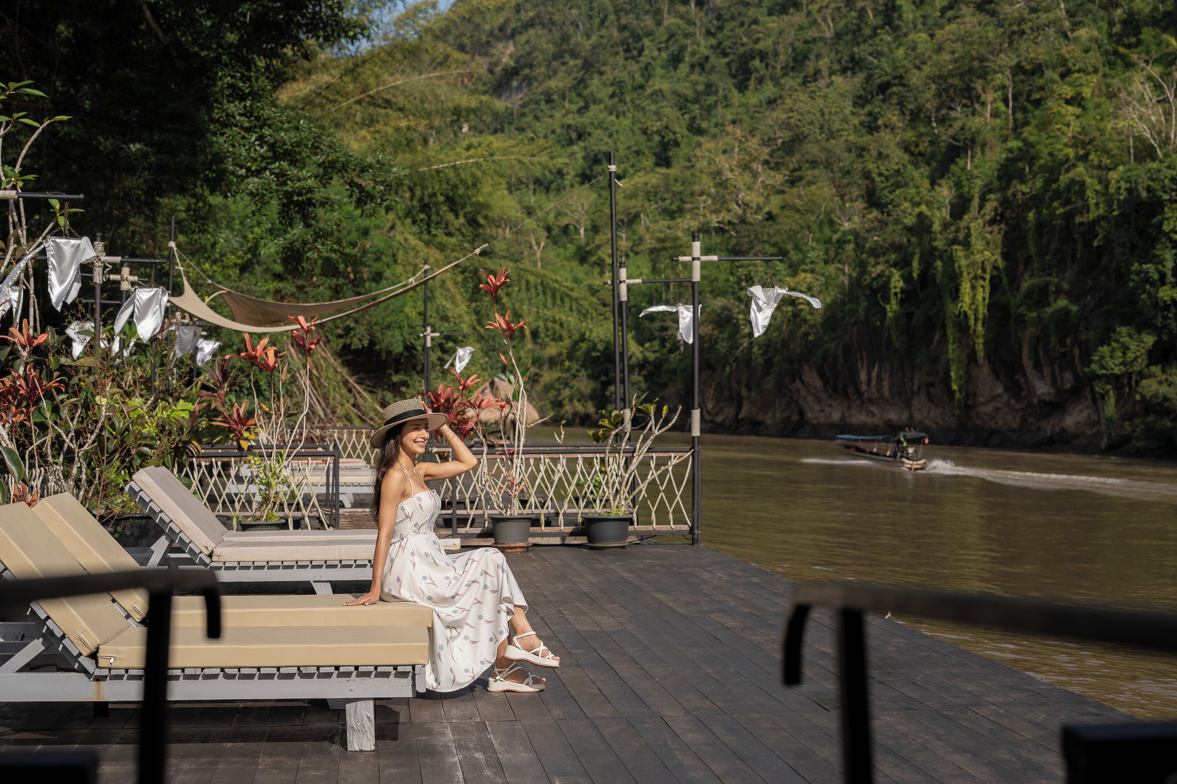 A woman sitting on a wooden deck with lounge chairs, overlooking a river with a boat in the distance.