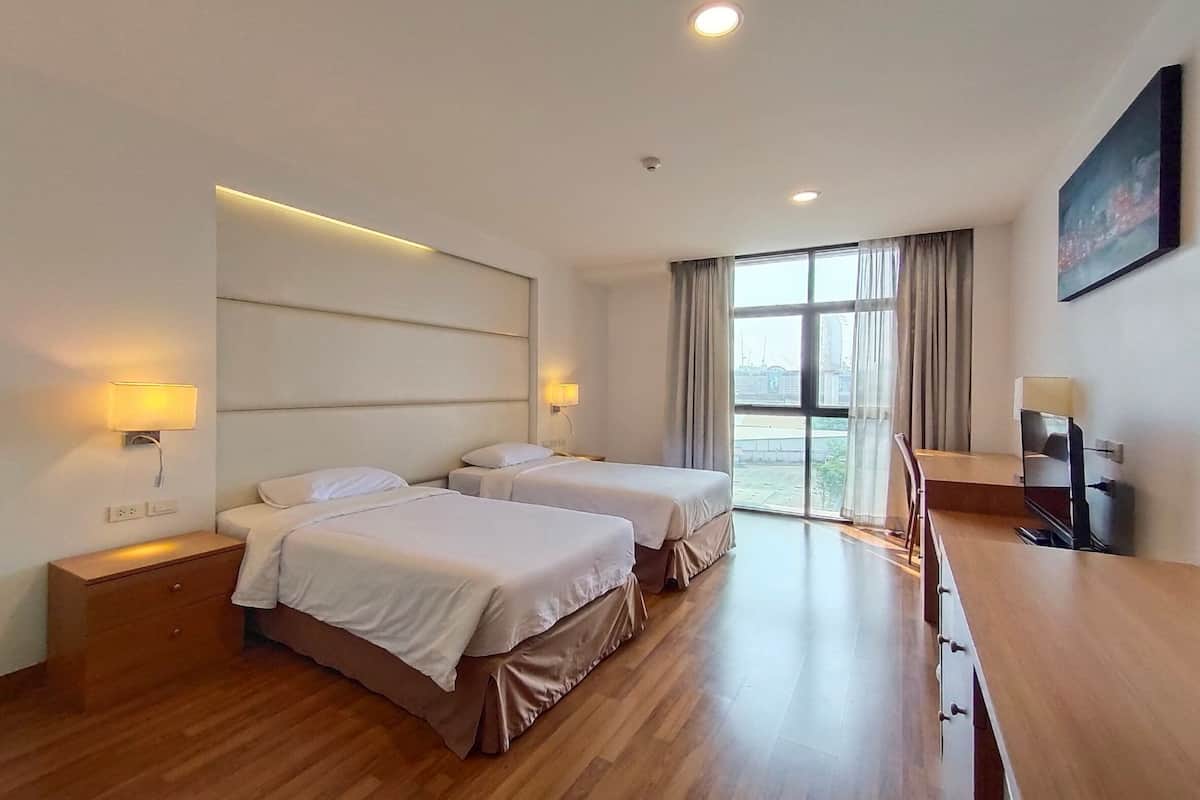 A hotel room with two beds, a wooden desk, a TV, and a large window with curtains.