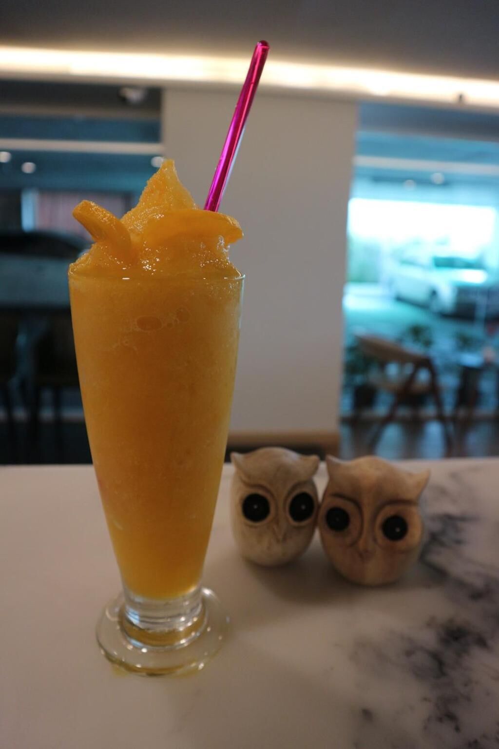Un grand verre rempli d’une boisson orange, avec une paille rose, posé sur une table avec deux statuettes de hiboux.