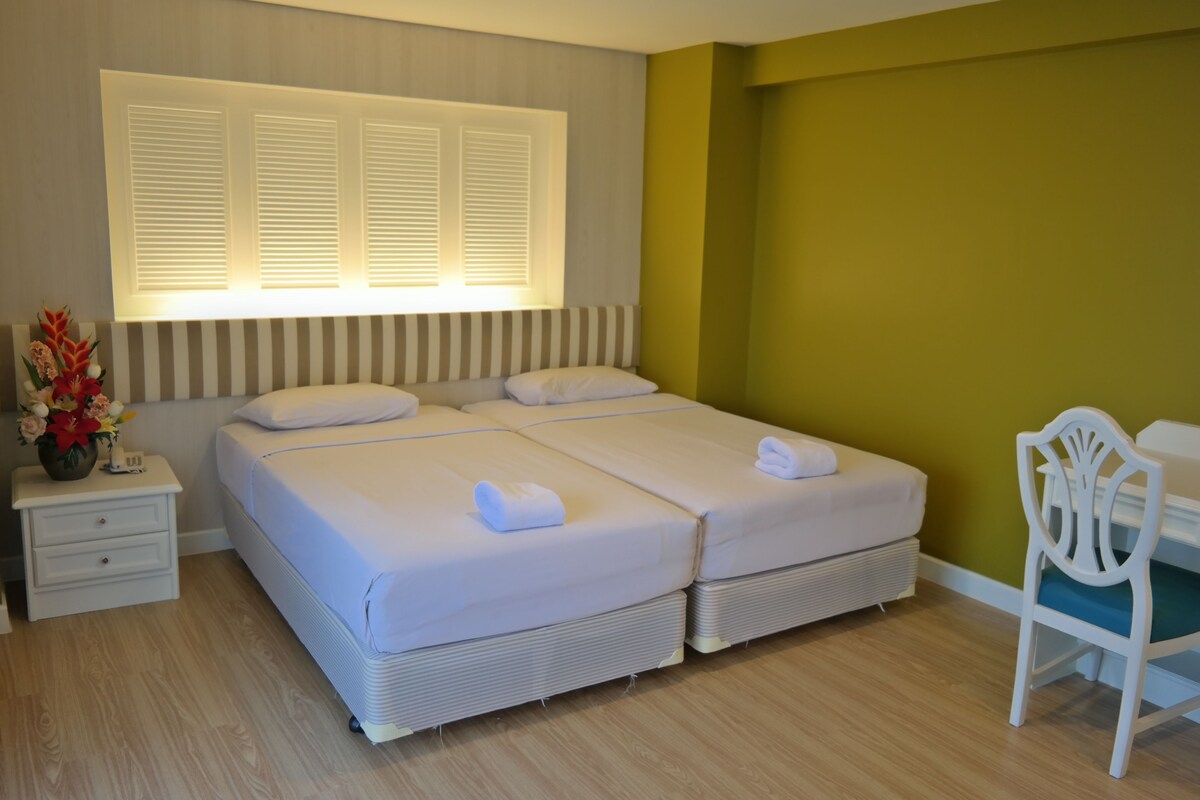 A double bed with white linens, a bedside table with a vase of flowers, and a chair with a white cushion.