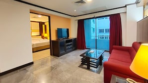 Quarto de hotel com uma cama, televisão, sofá vermelho, mesa de centro de vidro e porta de correr para a varanda.
