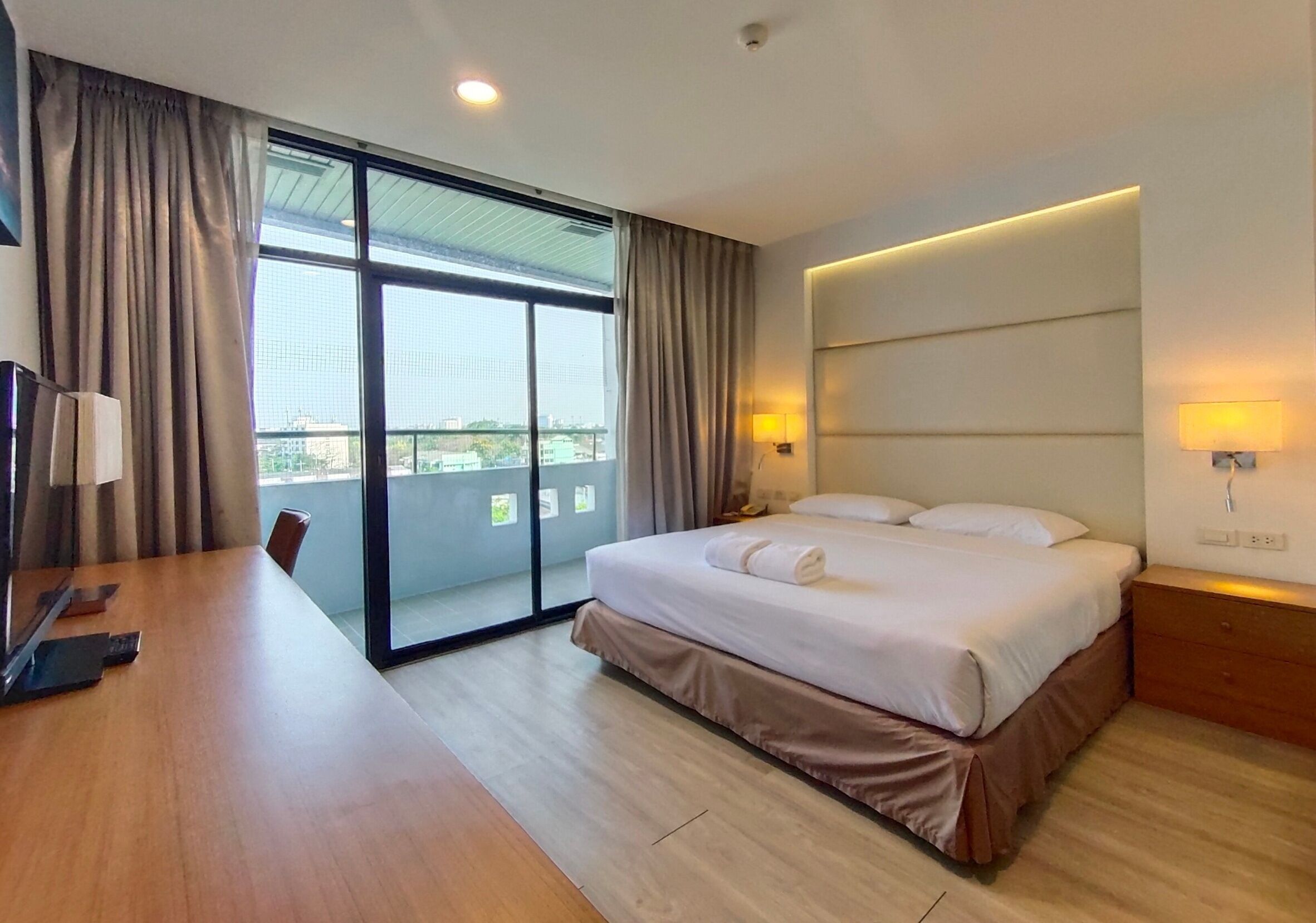 A hotel room with a large bed, a desk, and a view of the city through a sliding door.