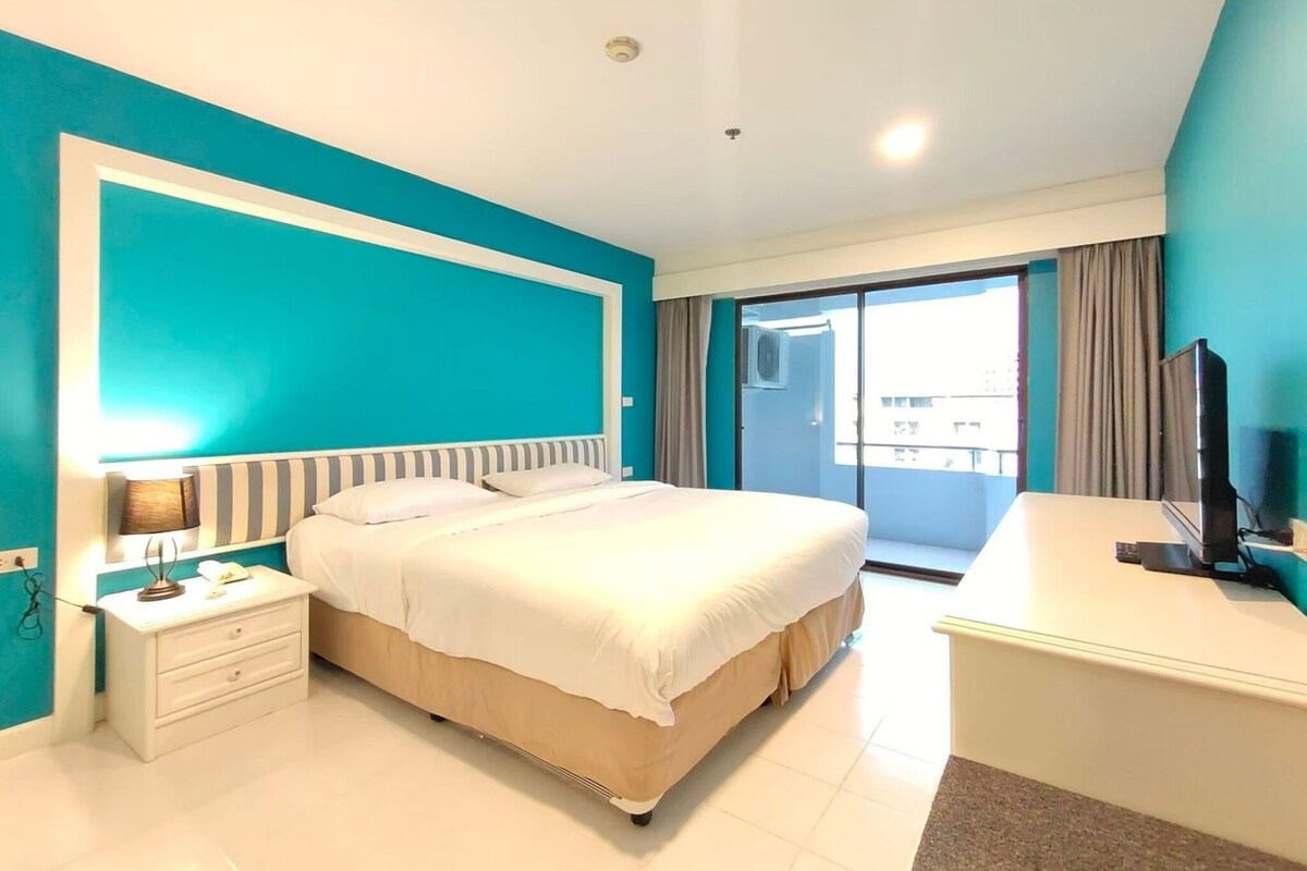 A hotel room with a bed, a desk, a TV, a nightstand, and a sliding glass door leading to a balcony.