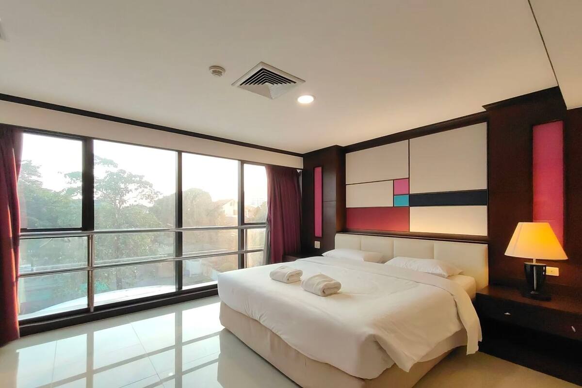 A modern hotel room with a large bed, a nightstand, a lamp, and a view of the outdoors through large windows.