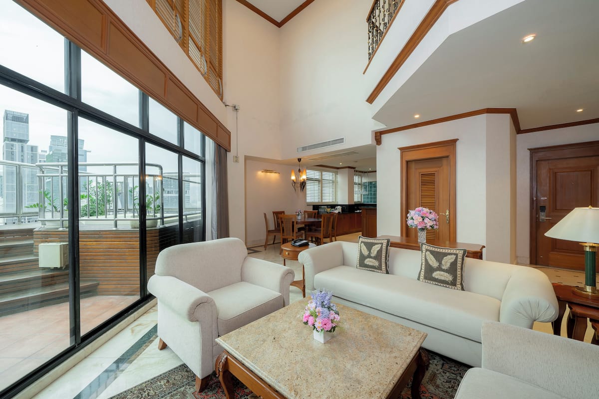 A spacious living area with a large window, a sofa, a coffee table with flowers, and a dining area in the background.
