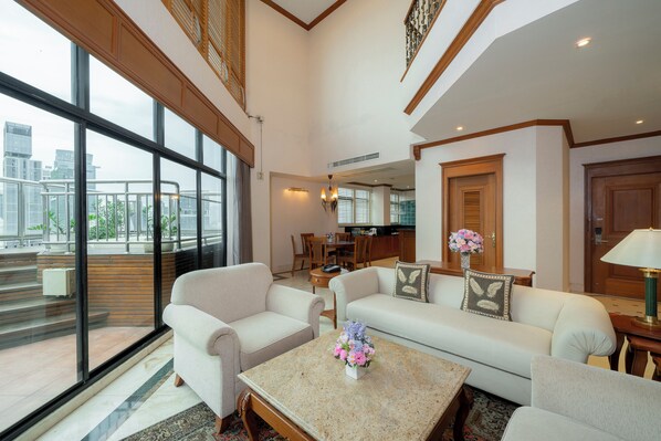 A spacious living area with a large window, a sofa, a coffee table with flowers, and a dining area in the background. - Grand Diamond Suite (Bangkok)