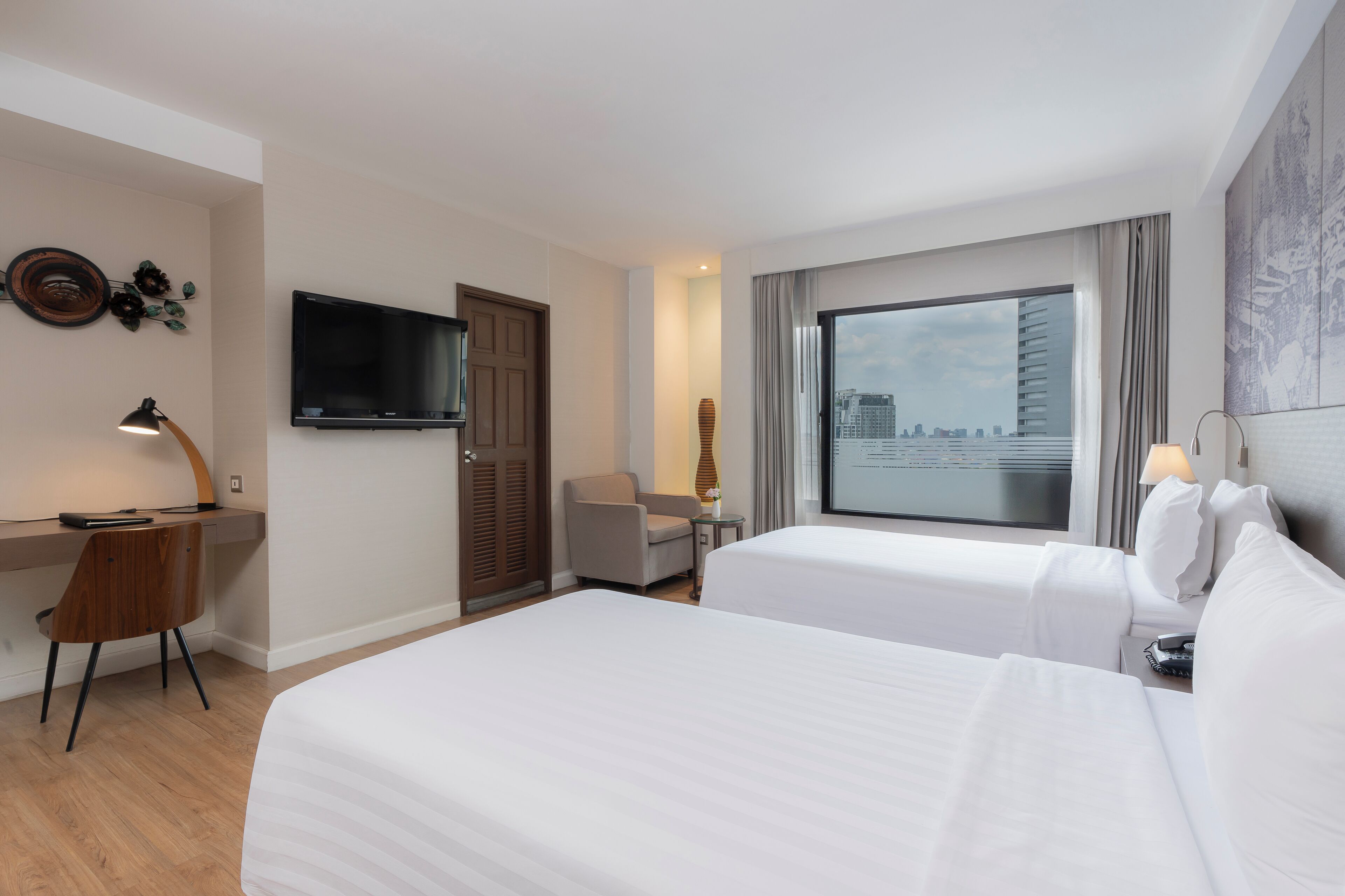 A hotel room with two beds, a desk, a chair, a TV, and a large window with a city view.