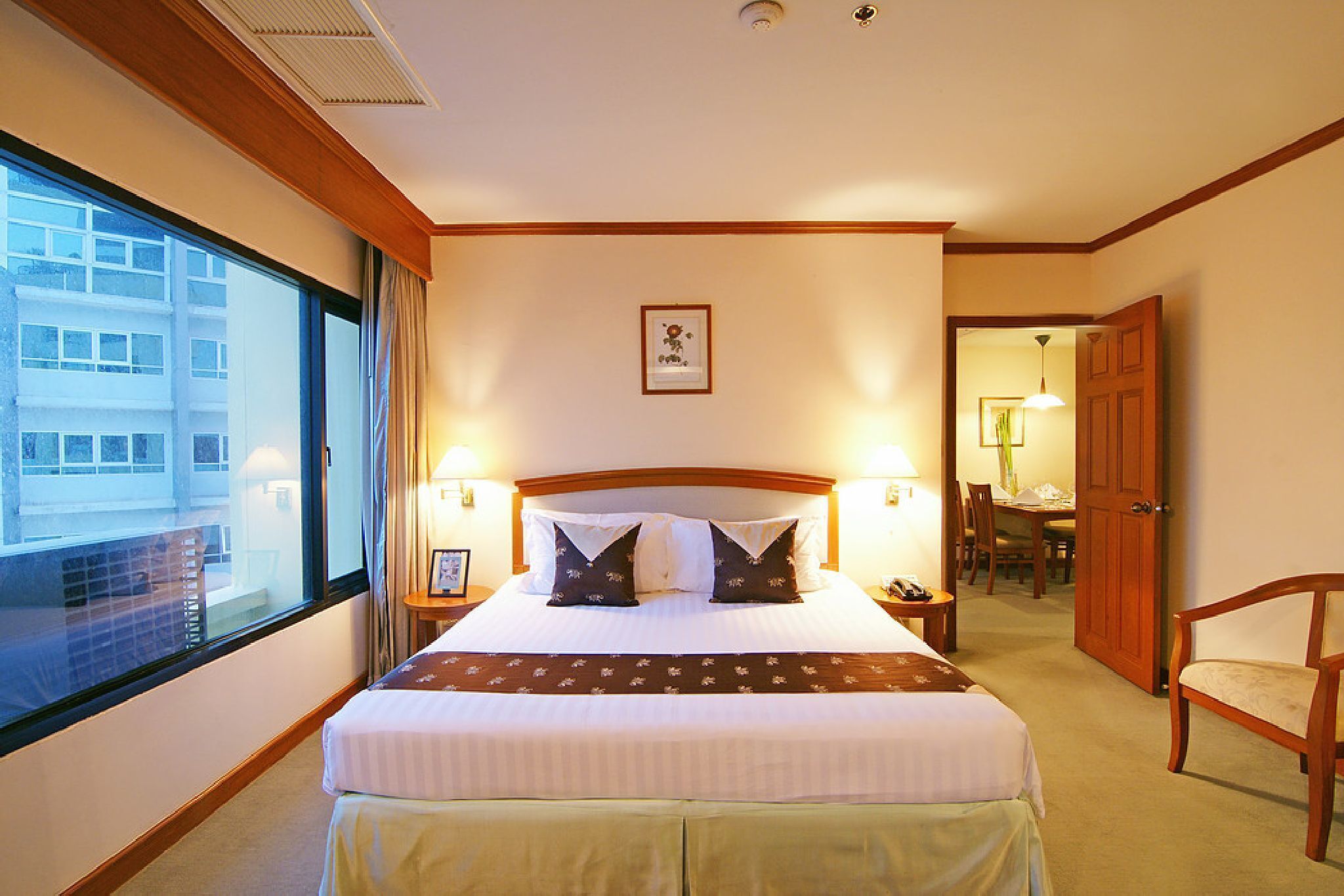 A hotel room with a large bed, two bedside lamps, a framed picture on the wall, a wooden door, and a view of a building outside.