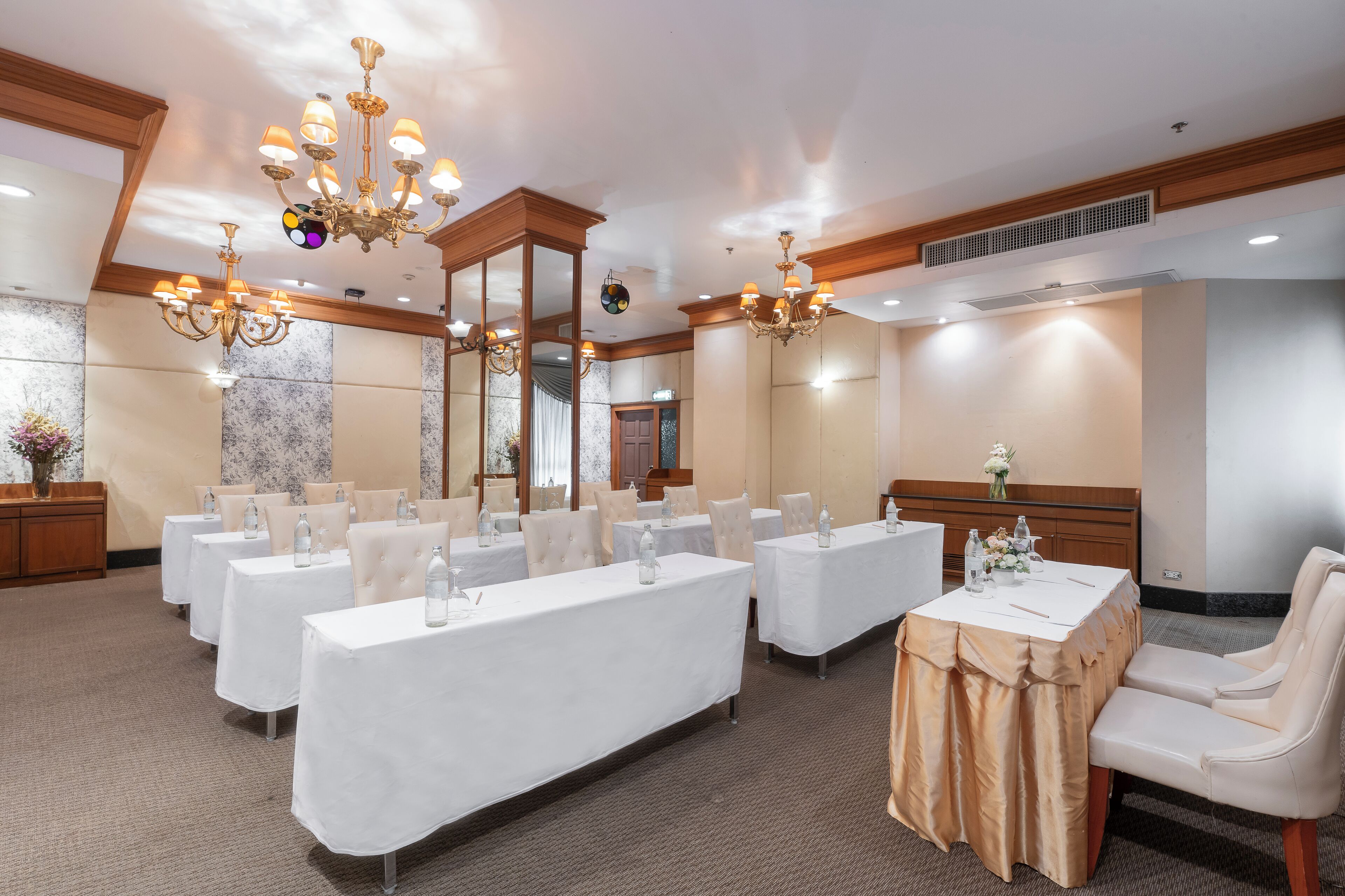 A hotel conference room with multiple tables, white chairs, chandeliers, and a wooden ceiling.