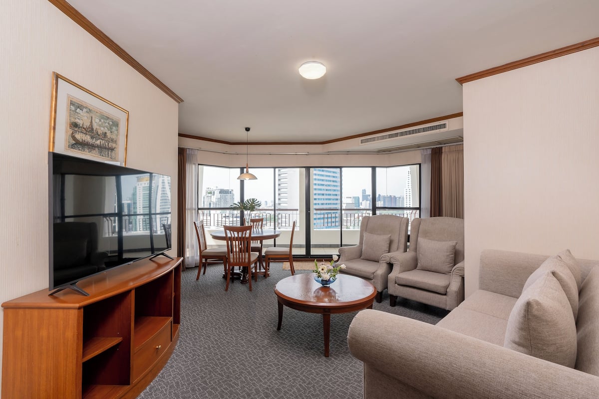 A hotel living room with a flat-screen TV, a sofa, a coffee table, and a dining area with chairs and a table.