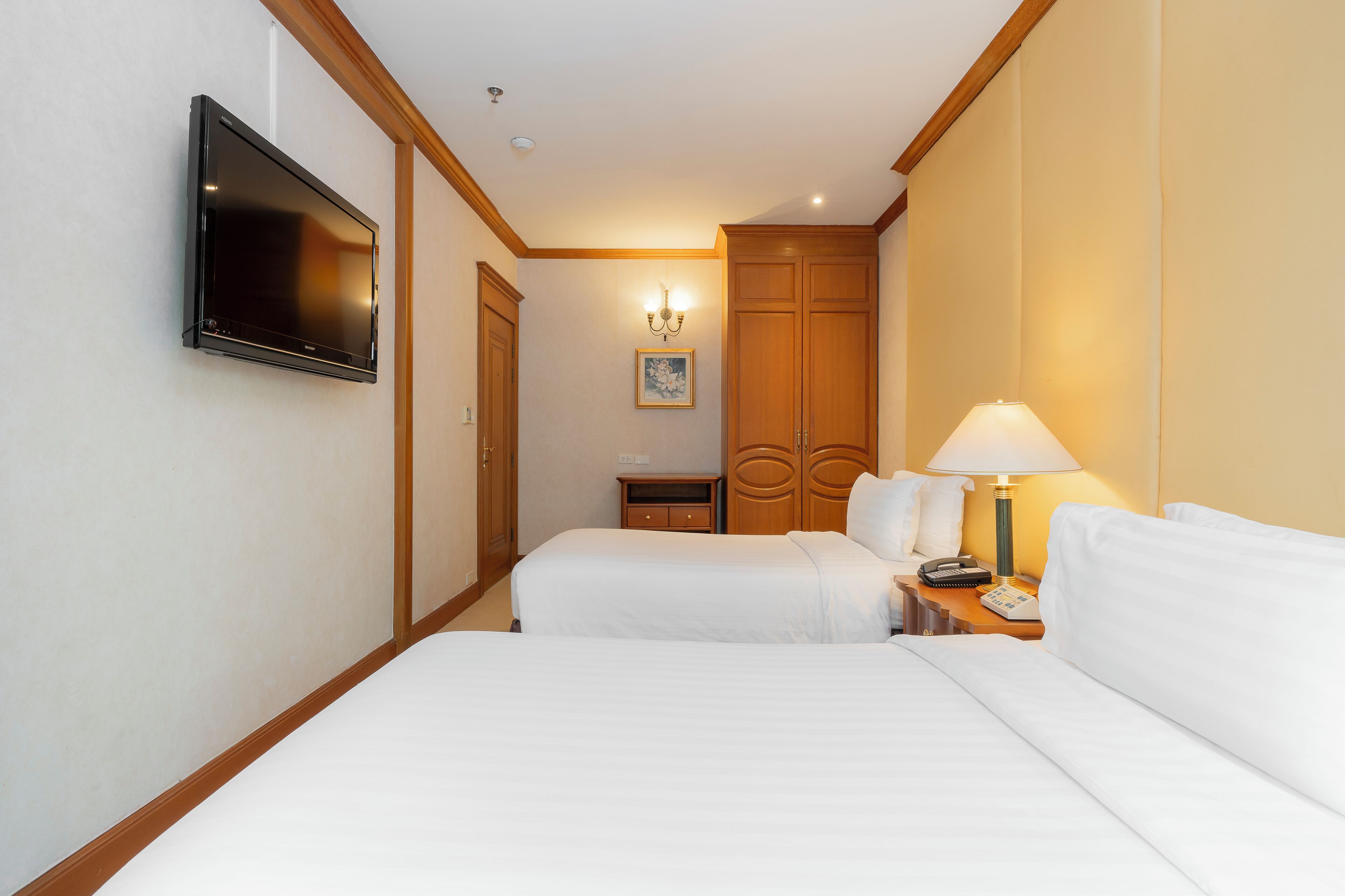 Une chambre d’hôtel avec deux lits, un téléviseur fixé au mur, une table de chevet avec une lampe et une armoire en bois.