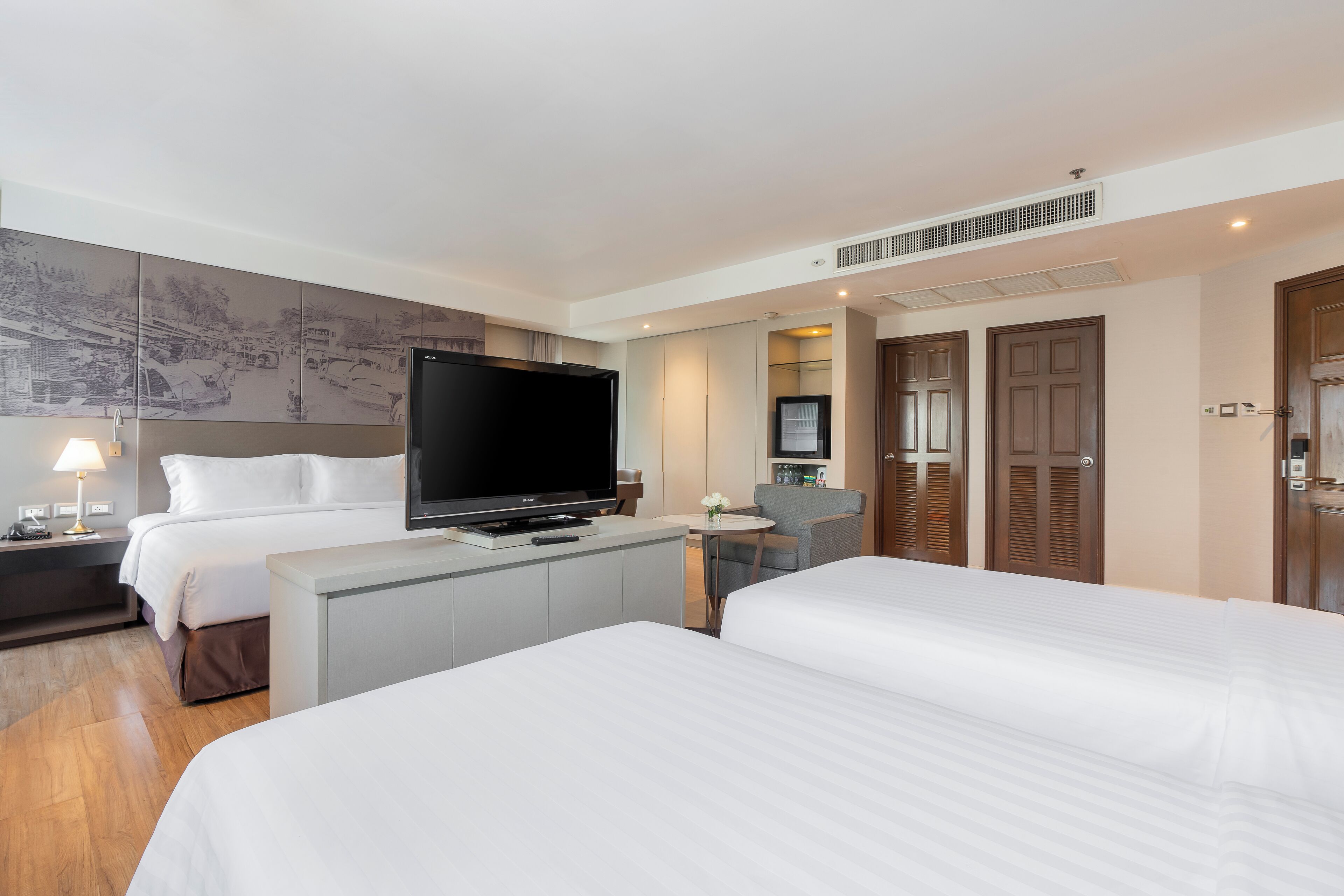 A hotel room with two beds, a flat-screen TV, a desk with a chair, a small table with a vase, and a wall-mounted TV.
