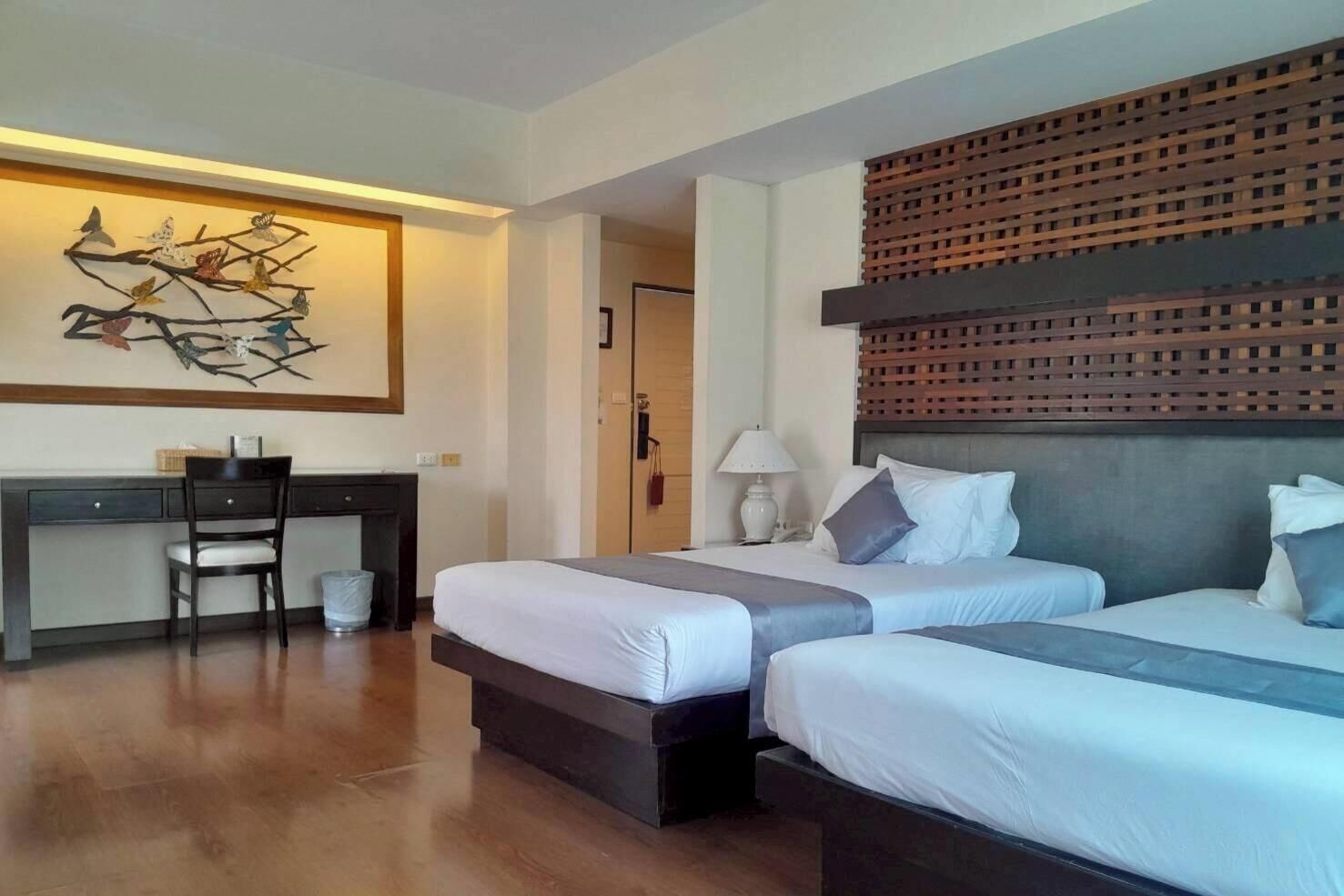 A hotel room with two beds, a desk, and a chair. There is a decorative wall piece above the desk and a wooden slat design on the wall behind the beds.