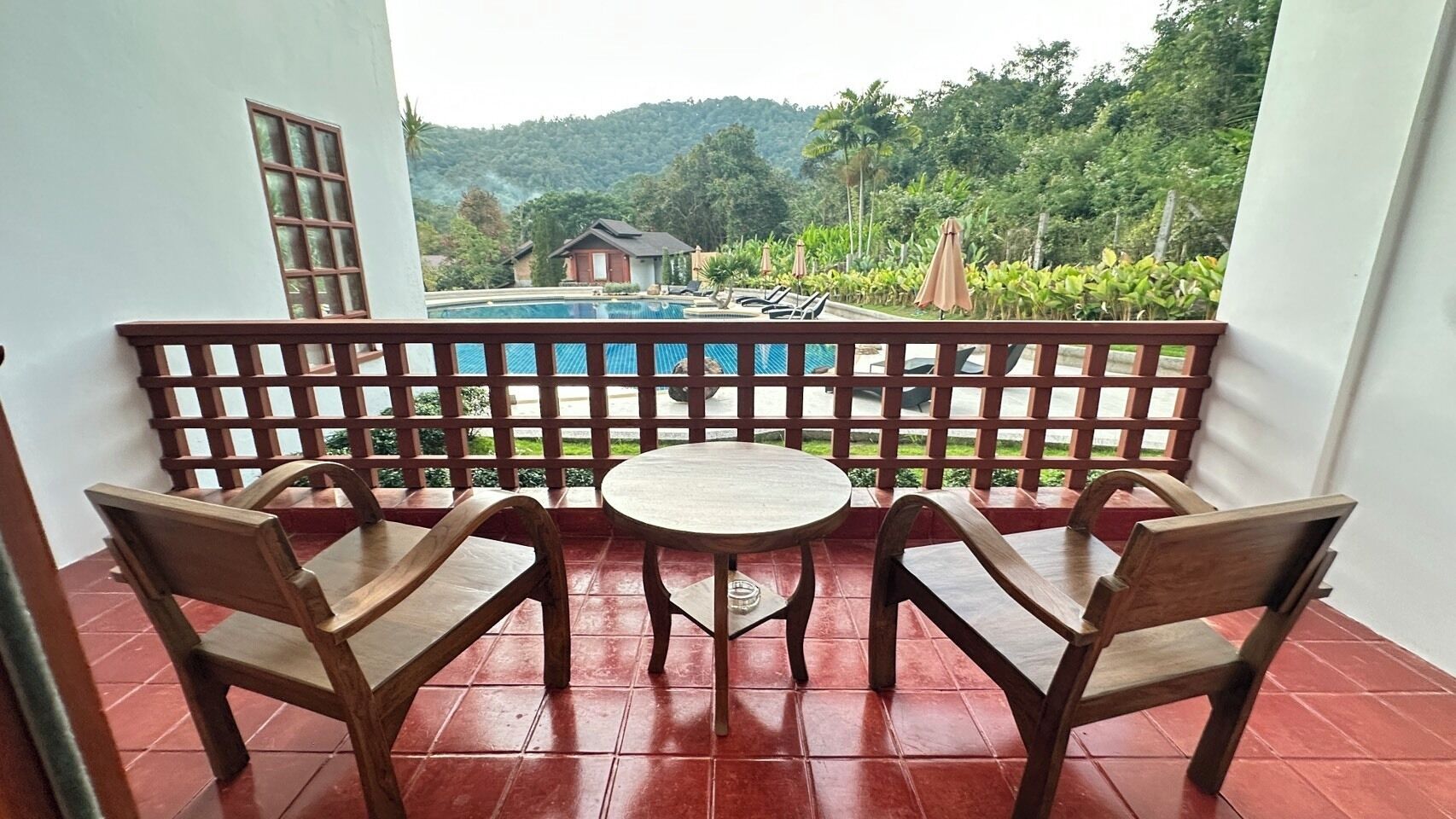 A balcony with two wooden chairs and a small round table, overlooking a swimming pool and lush greenery.