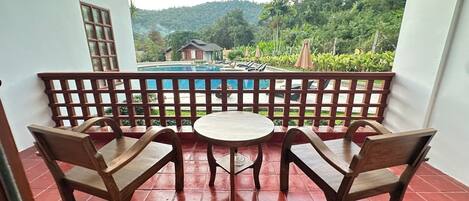 A balcony with two wooden chairs and a small round table, overlooking a swimming pool and lush greenery.