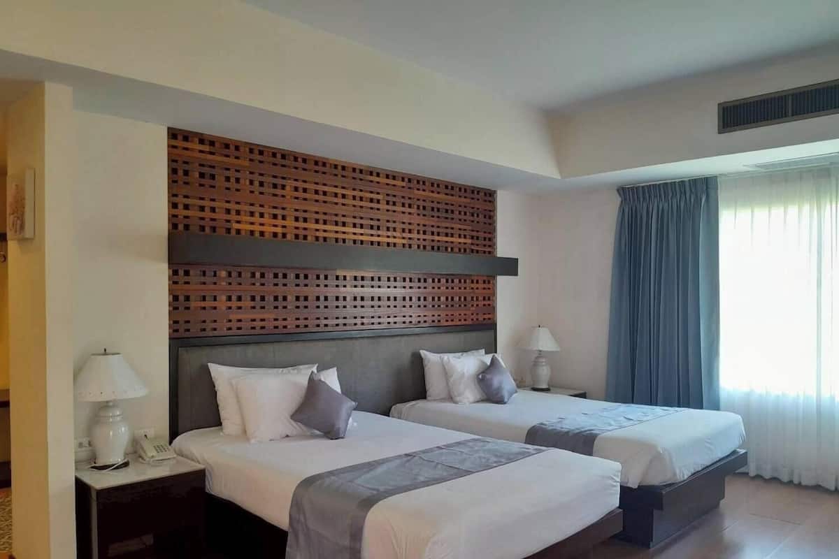 Une chambre d’hôtel avec deux lits, une tête de lit en bois, une table de chevet avec une lampe et une fenêtre avec des rideaux.