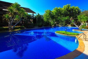 Une piscine dont le bord est recouvert de carreaux, entourée de plantes tropicales et avec un bâtiment visible au loin.