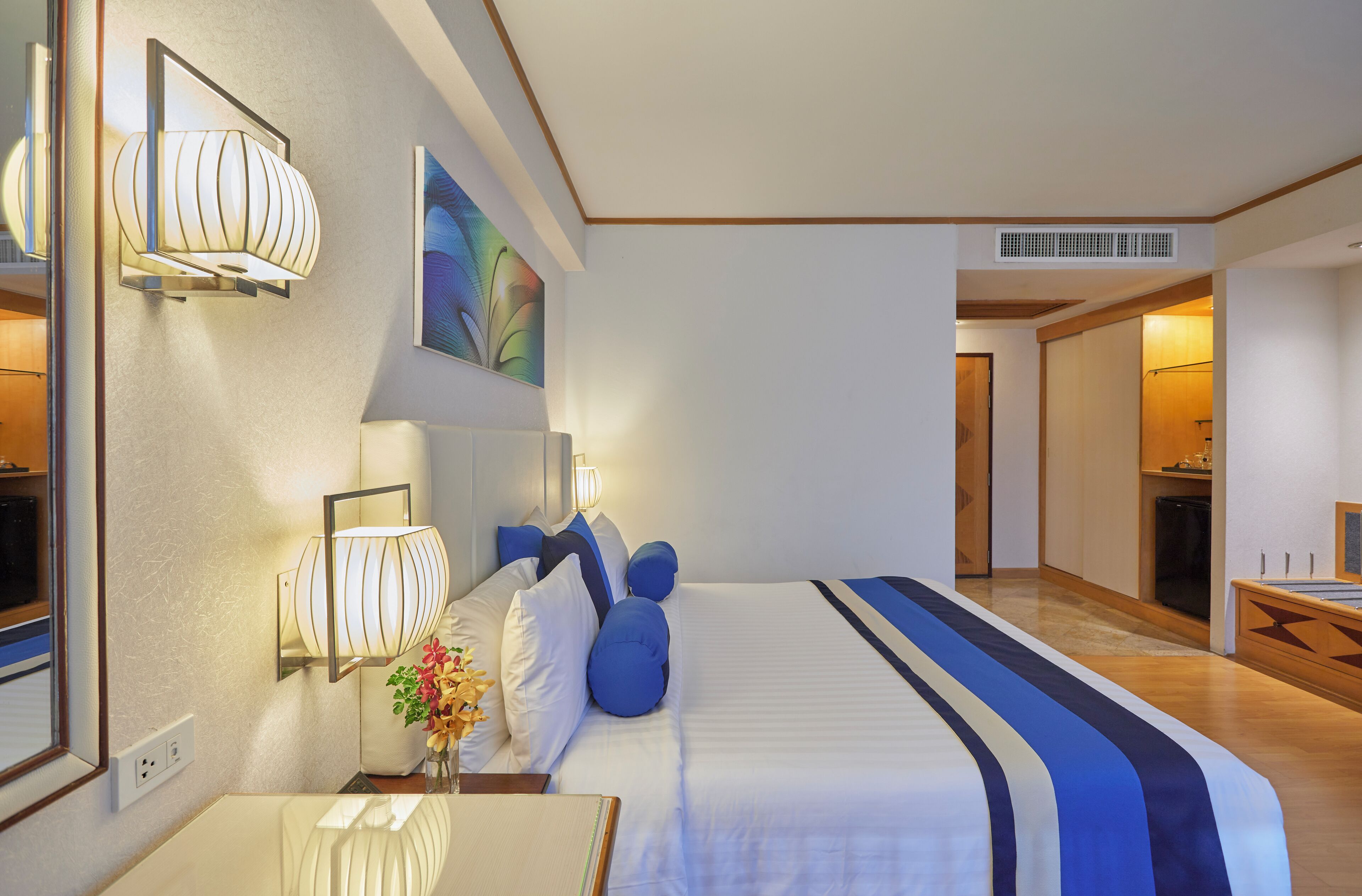 Ein Hotelzimmer mit einem großen Bett, Nachttischlampen, einem Spiegel, einem Bild an der Wand und Blick in das angrenzende Zimmer.