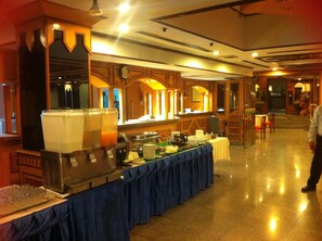 A buffet setup with multiple serving stations, a clock on the wall, and a person walking in the background. - Inn Come Hotel Chiangrai (Chiang Rai)