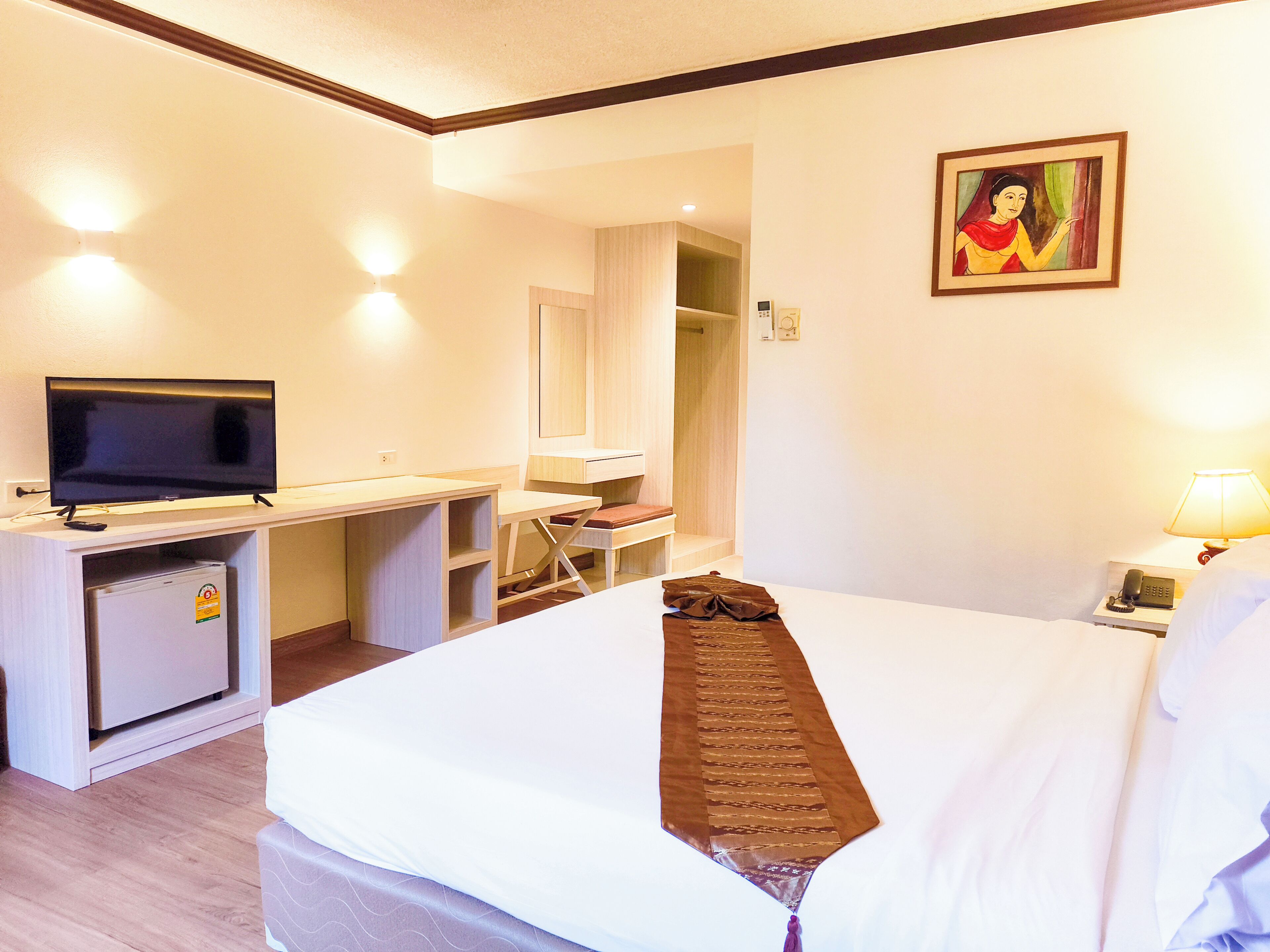 Een hotelkamer met een bed, een bureau met een televisie, een kleine koelkast, een stoel, een nachtkastje met een lamp en een schilderij aan de muur.