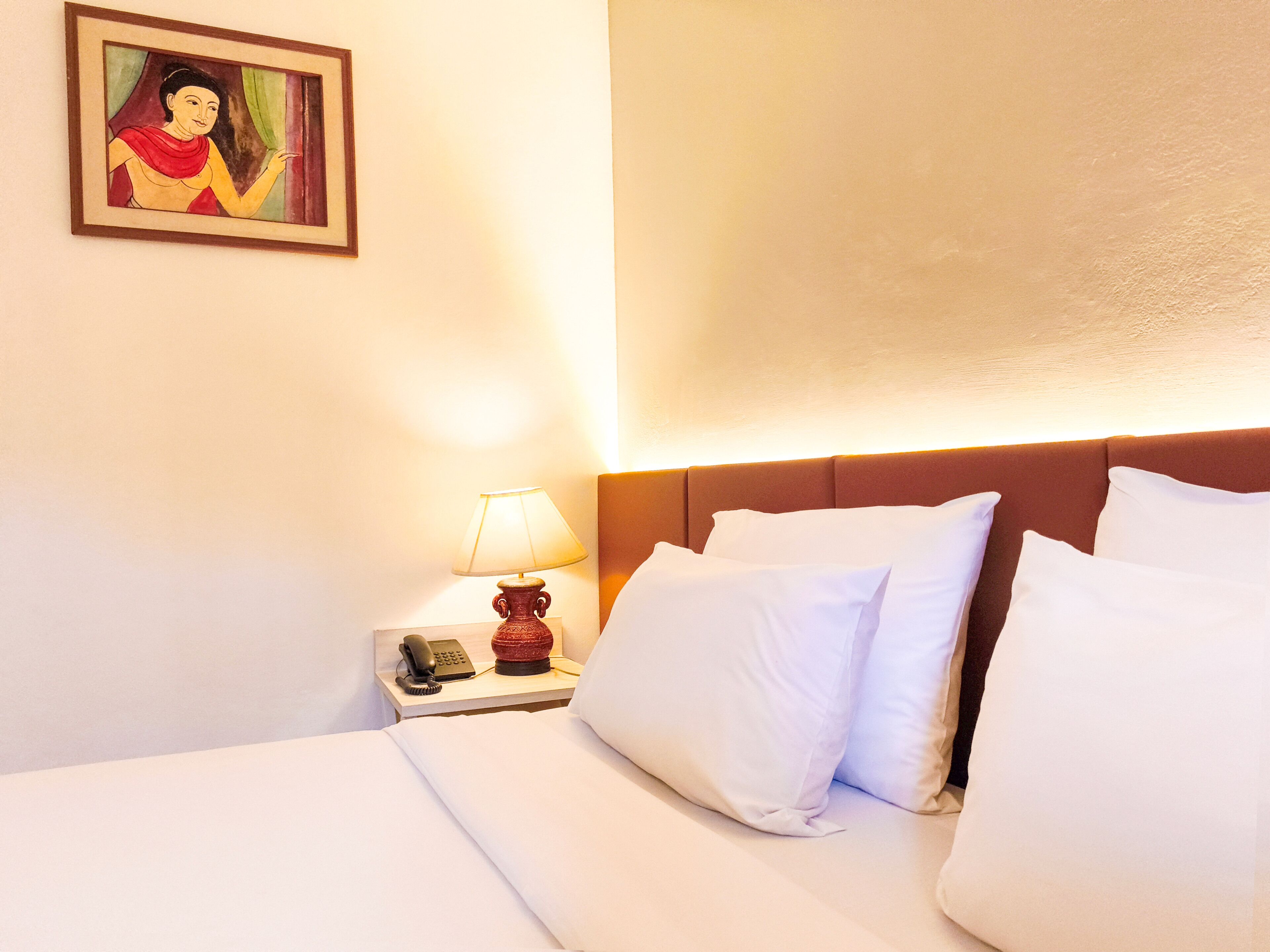 A hotel room with a bed, white pillows, a nightstand with a lamp, a phone, and a painting on the wall.