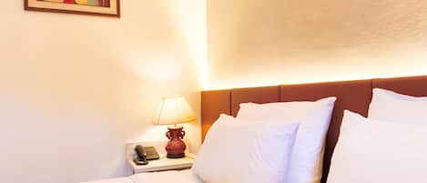 A hotel room with a bed, white pillows, a nightstand with a lamp, a phone, and a painting on the wall.