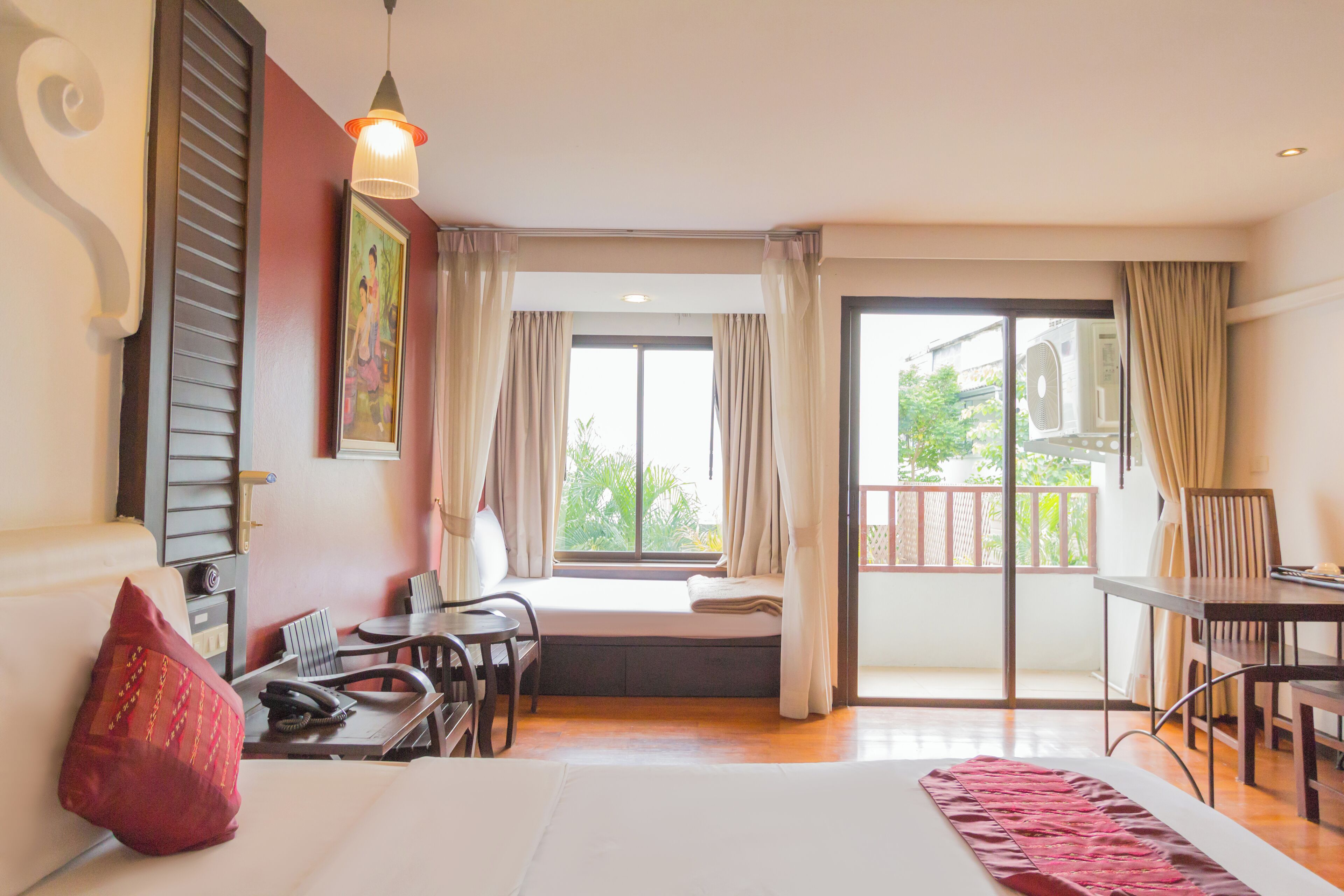 A hotel room with a bed, a desk, a chair, a window with curtains, and a balcony with a view of greenery.