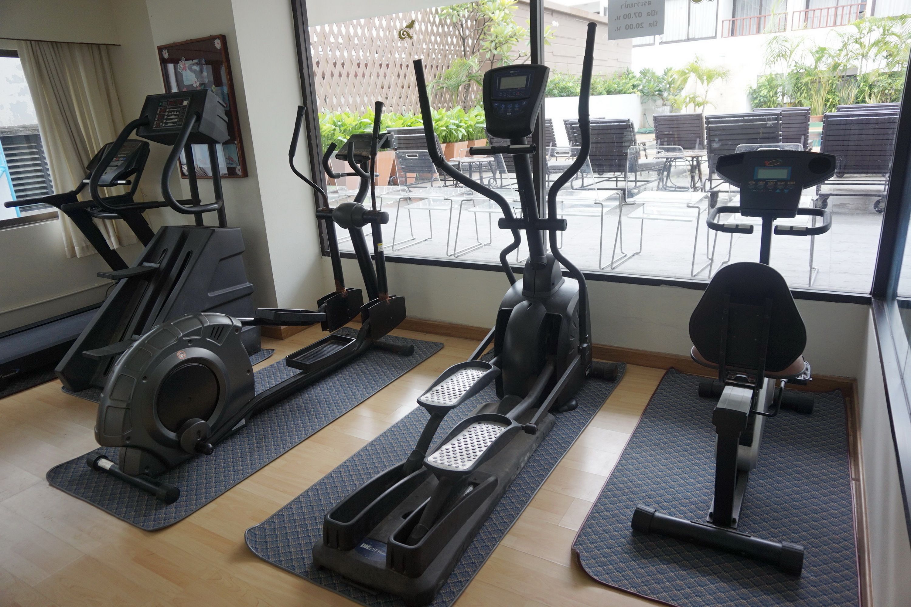 Un gimnasio con diferentes máquinas de cardio, colchonetas y vistas a una zona de estar exterior.