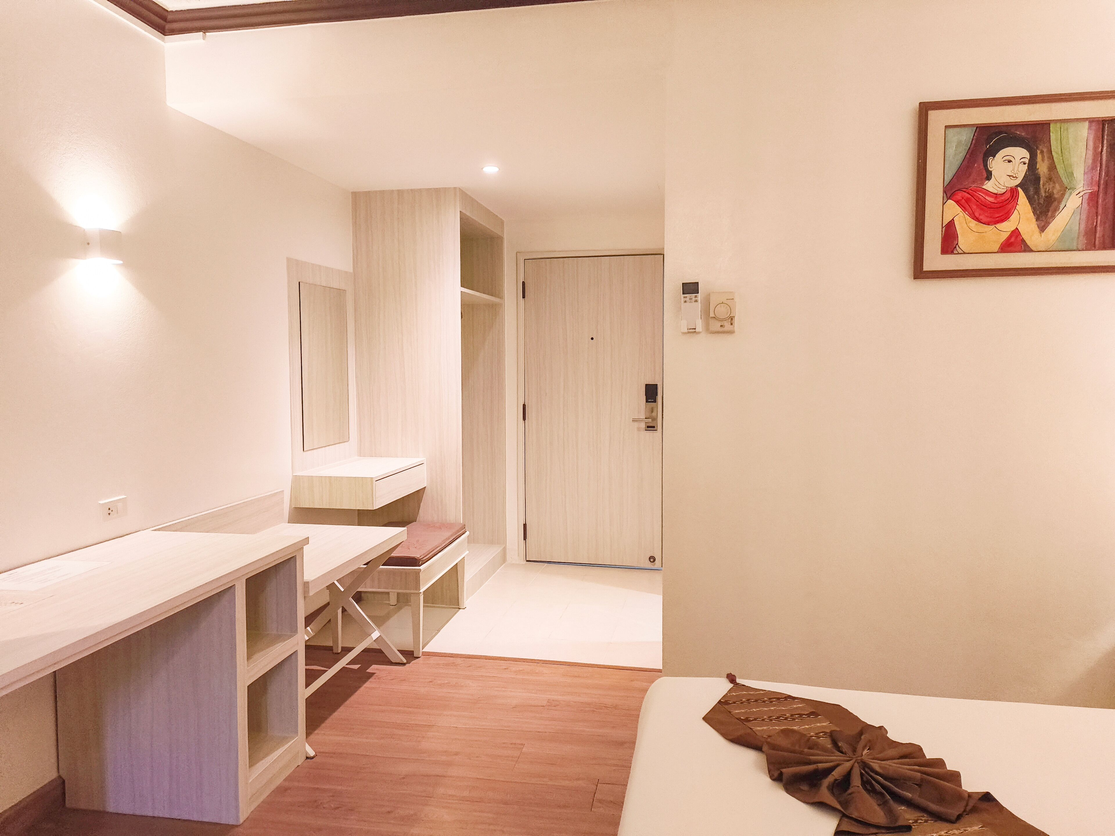 A modern hotel room with a wooden desk, a bed, a wardrobe, and a painting on the wall.
