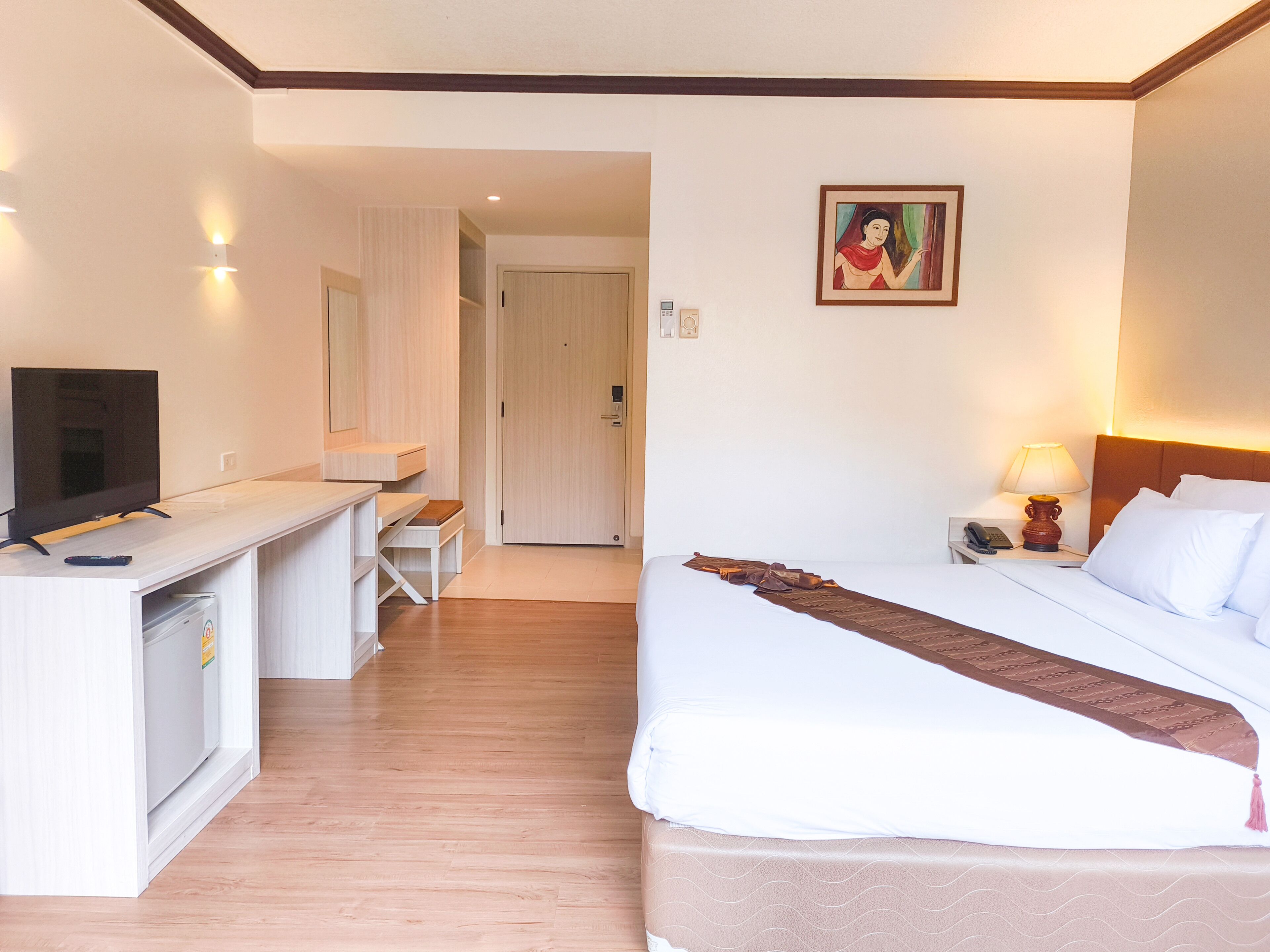 A hotel room with a bed, a desk with a TV, a chair, a door, and a painting on the wall.