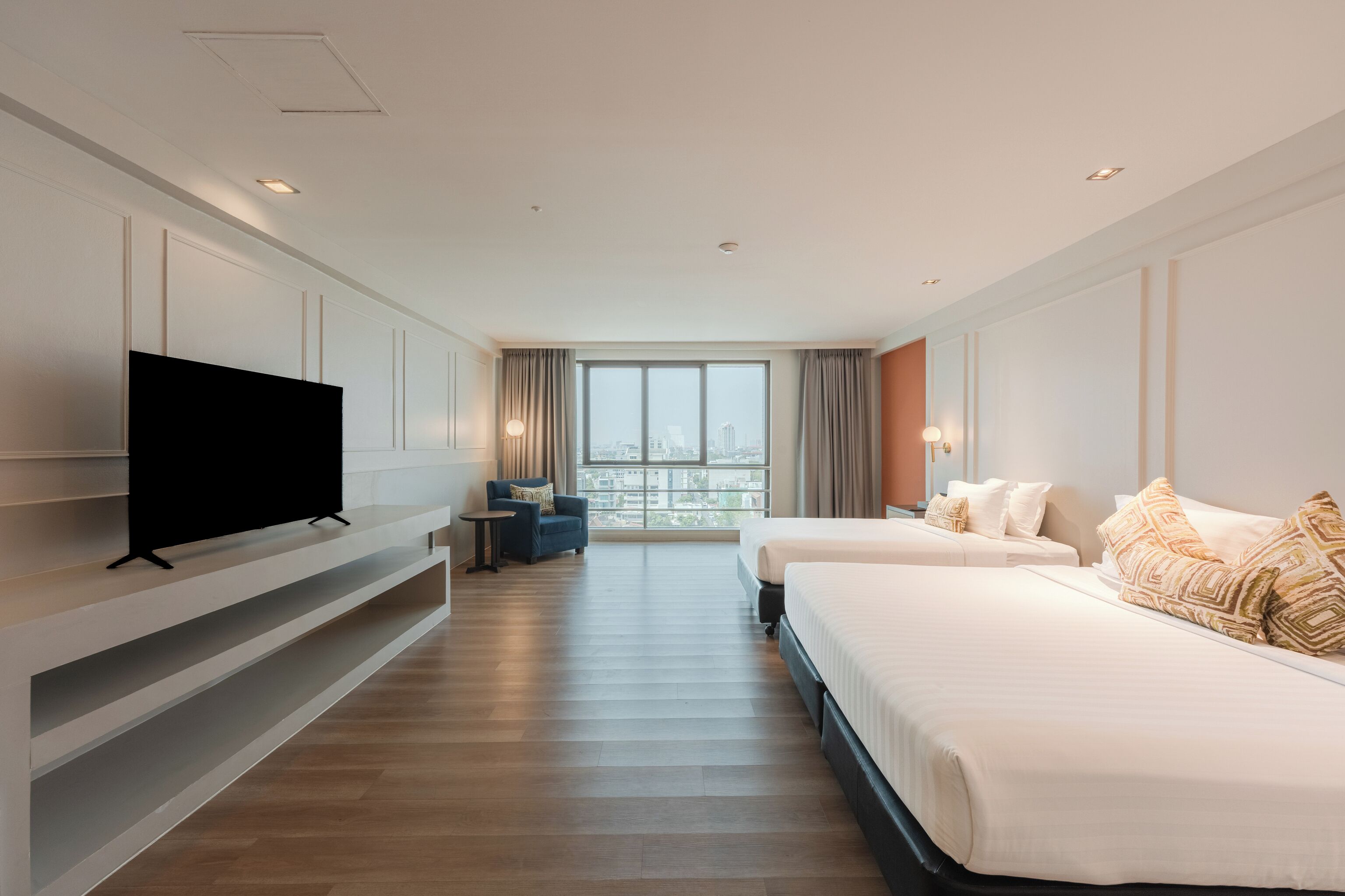 A hotel room with a TV, two beds, a sofa, a window with a city view, and a wooden floor.