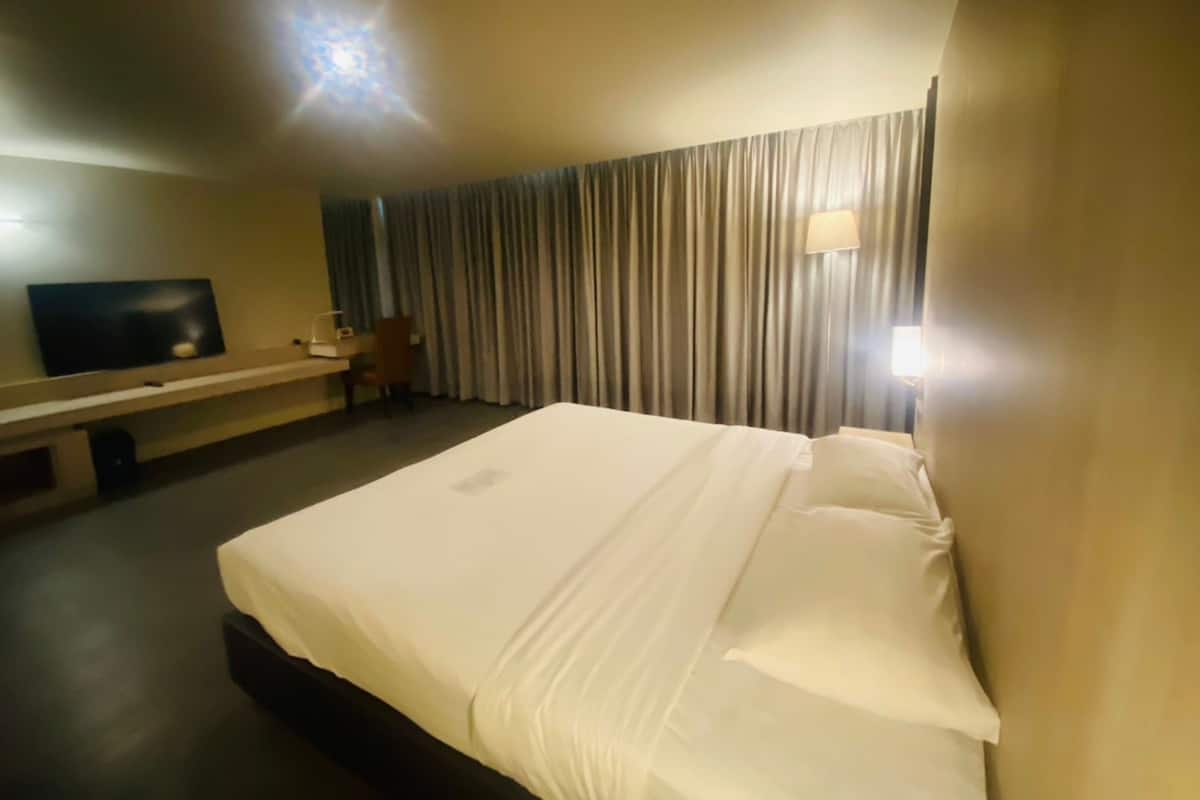 A hotel room with a large bed, a desk with a chair, a flat-screen TV on the wall, and a window with curtains.