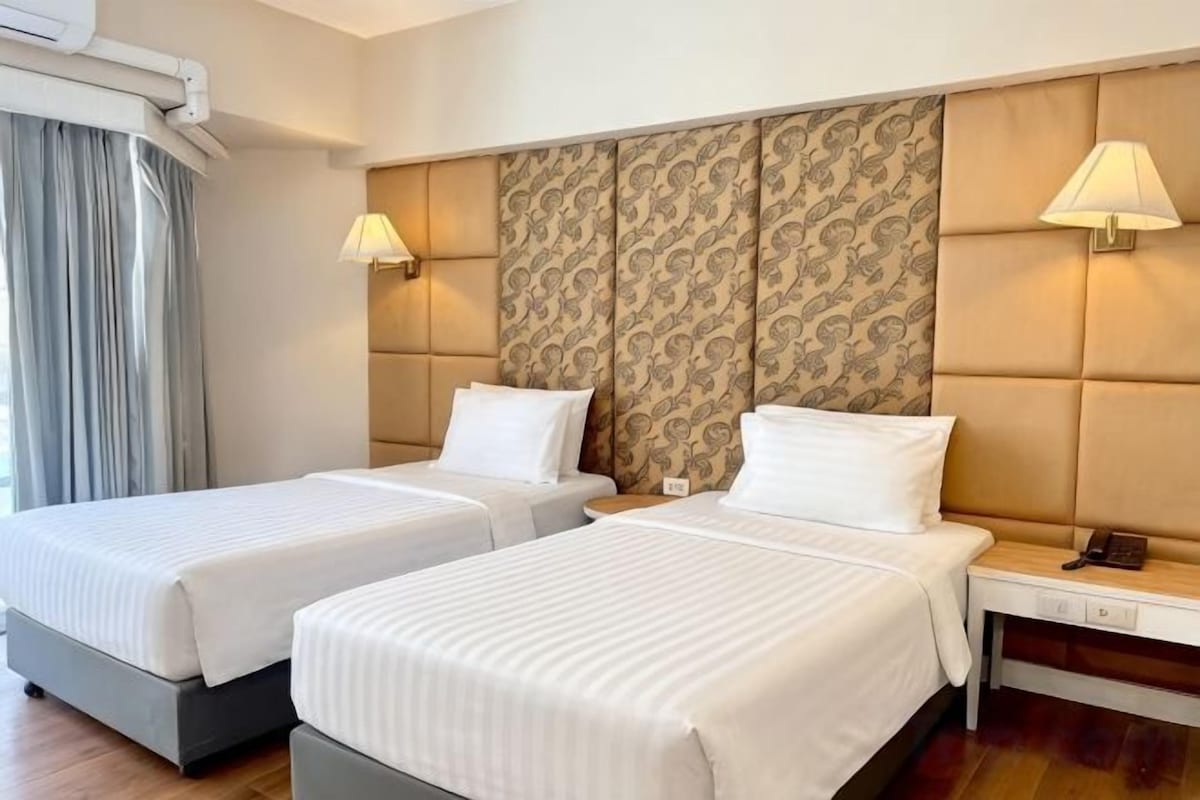 Habitació d'hotel amb dos llits, una taula de nit amb una làmpada i un capçal de llit estampat.