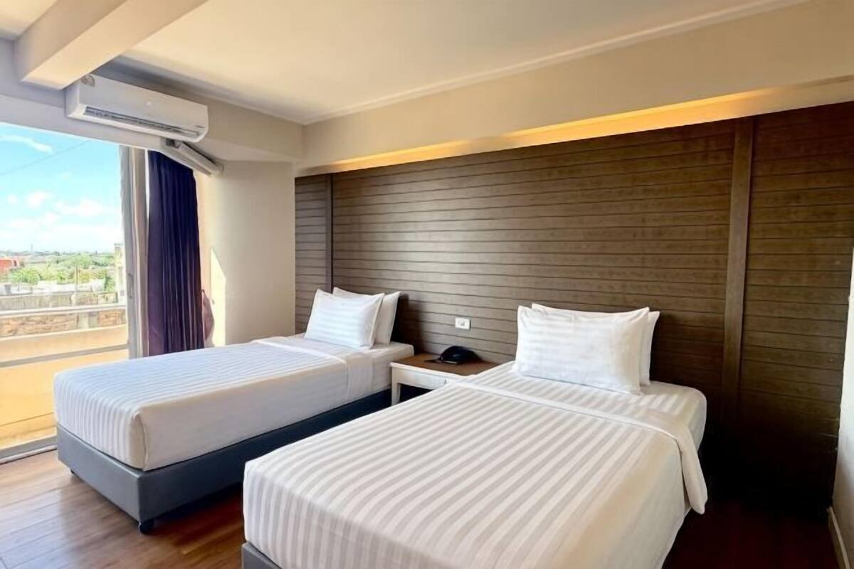 A hotel room with two beds, a wooden headboard, a nightstand, and a window with a view.