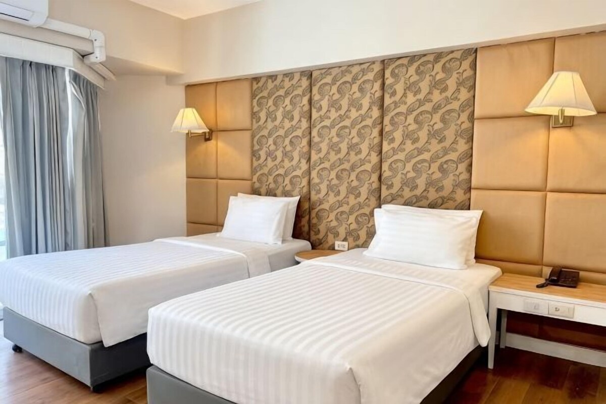 A hotel room with two beds, a bedside table with a lamp, a headboard with a patterned design, and a window with curtains.