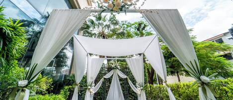 An outdoor wedding arch with white drapery and floral decorations, set against a backdrop of lush greenery and a modern building.