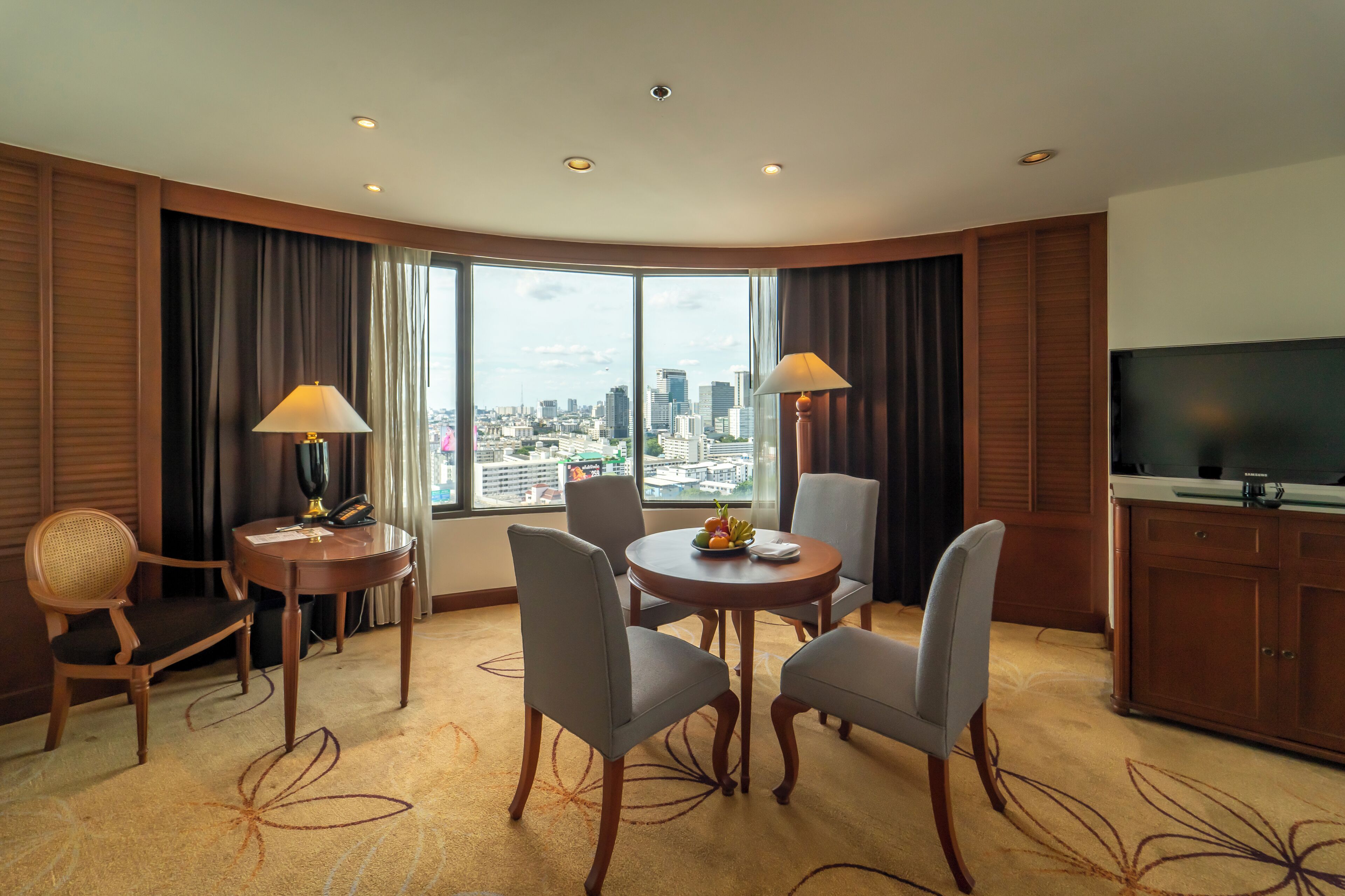 Habitación de hotel con un gran ventanal, una mesa de comedor con sillas, un televisor y un minibar.