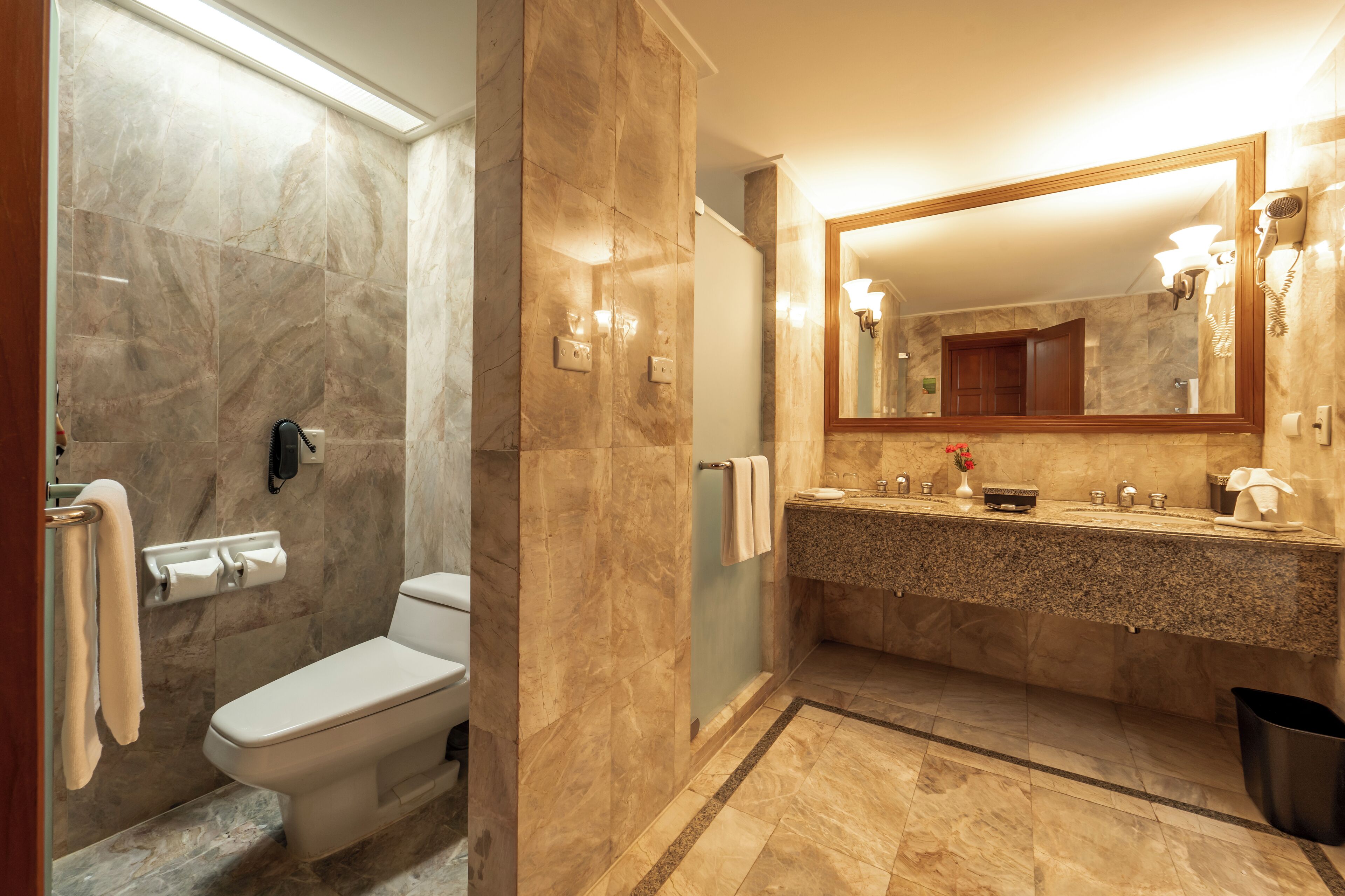 A bathroom with a marble shower, a toilet, a sink with a mirror, and a trash bin.