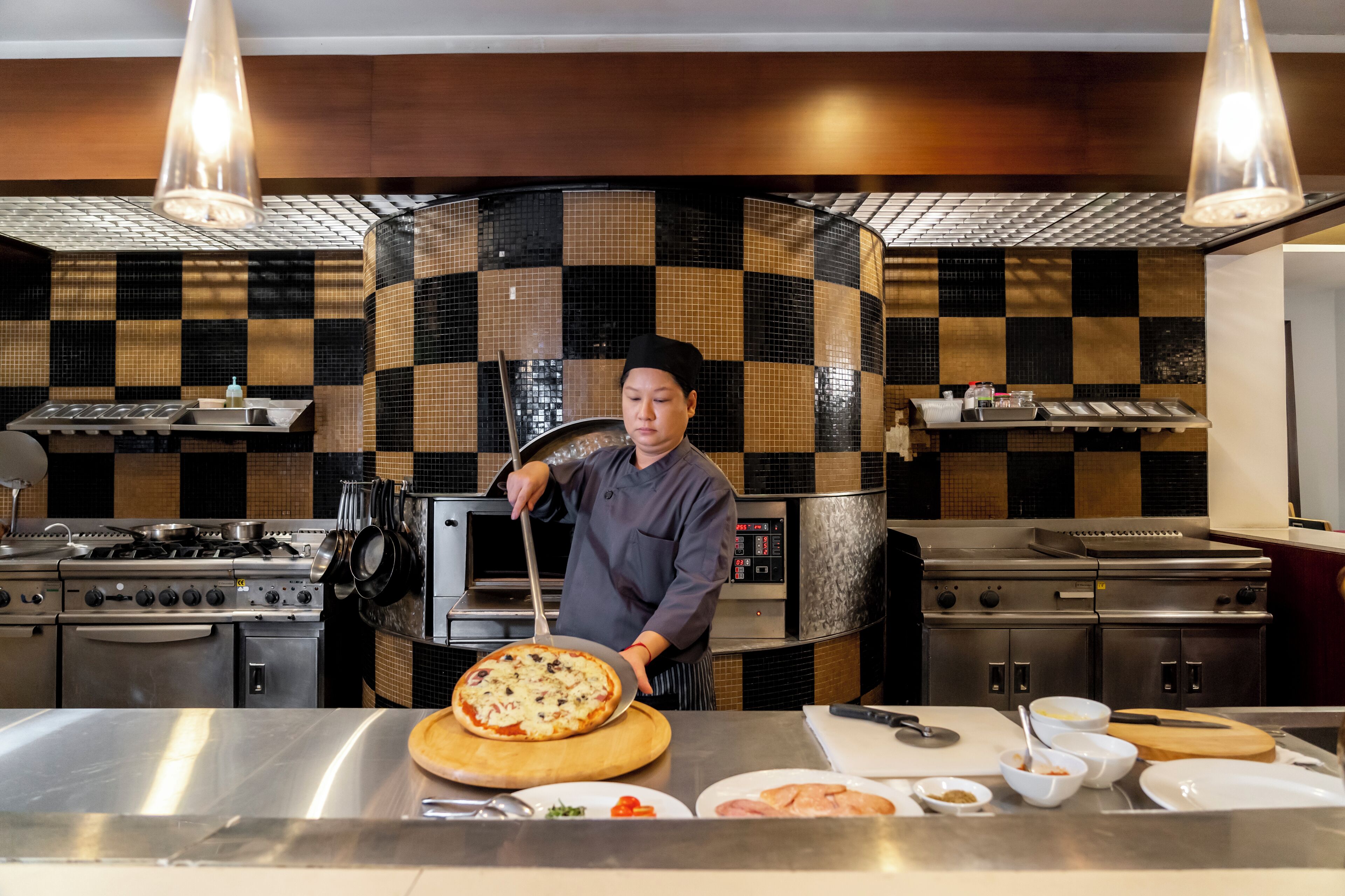 A chef in a modern kitchen with checkered tile backsplash, stainless steel appliances, and a pizza on a wooden board.