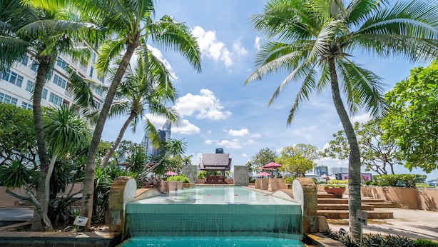 A swimming pool with a tiled edge, surrounded by palm trees and a building with a pagoda-style roof in the background. - Century Park Hotel (Bangkok)