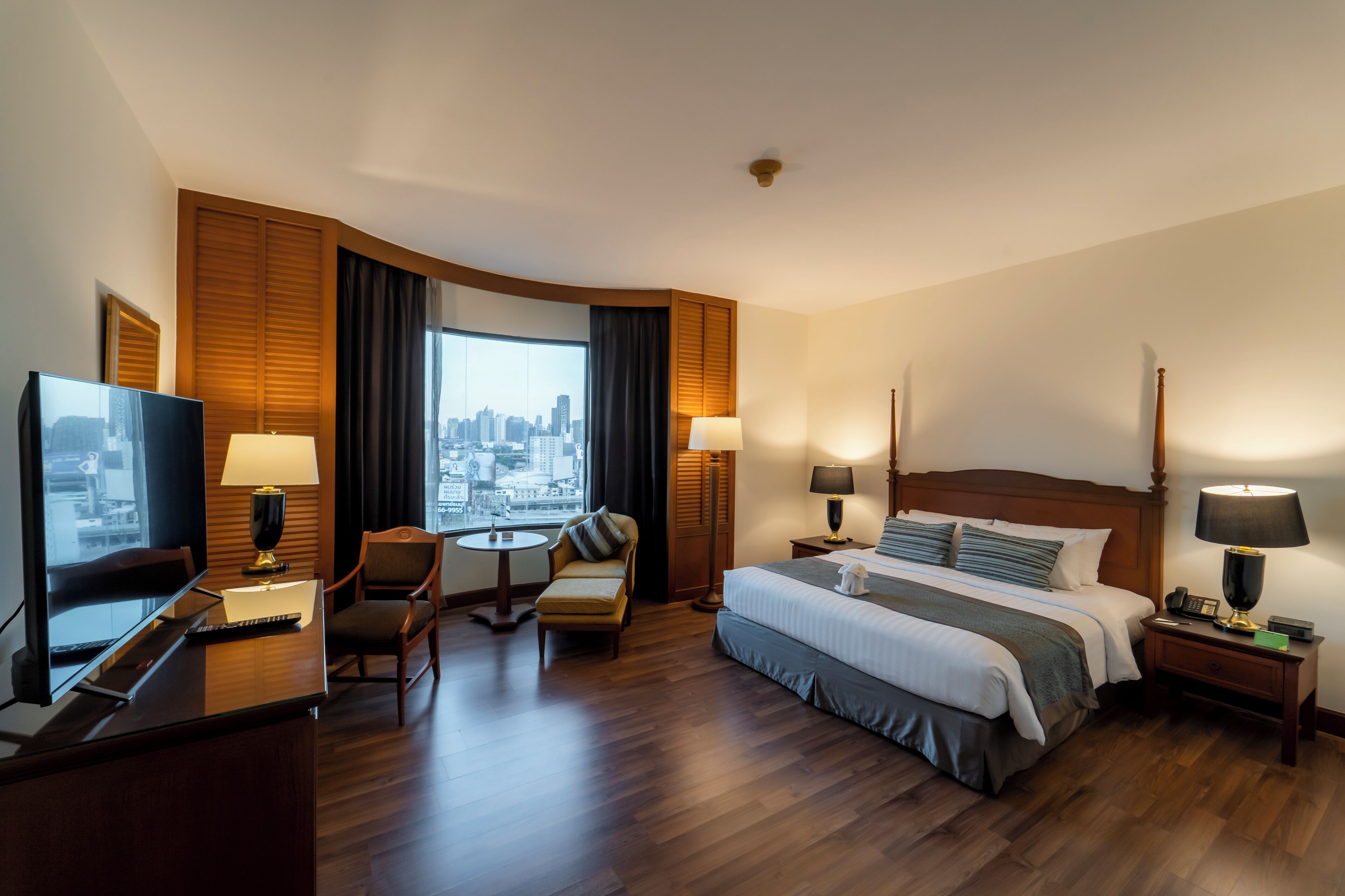 A hotel room with a large bed, a desk with a TV, a chair, a small table, and a view of a city skyline.