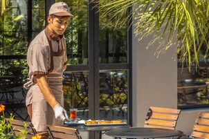 Una persona con uniforme sirve comida en una mesa al aire libre con sillas y un vaso de bebida.